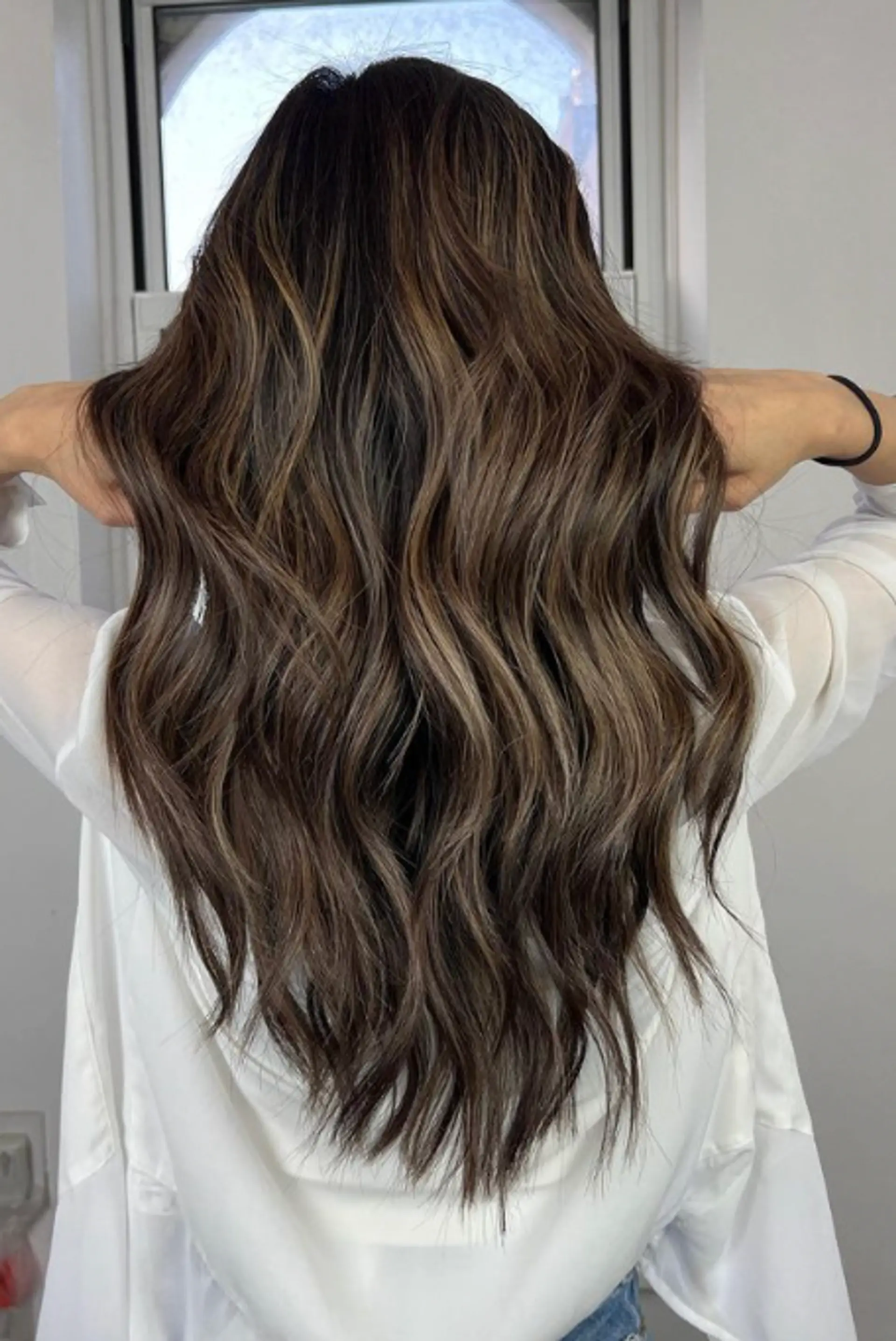 The style features long, cascading waves with a rich brown base color and ash blonde balayage highlights, creating a sun-kissed and multi-tonal effect. The texture is wavy, adding volume and movement to the look. 1