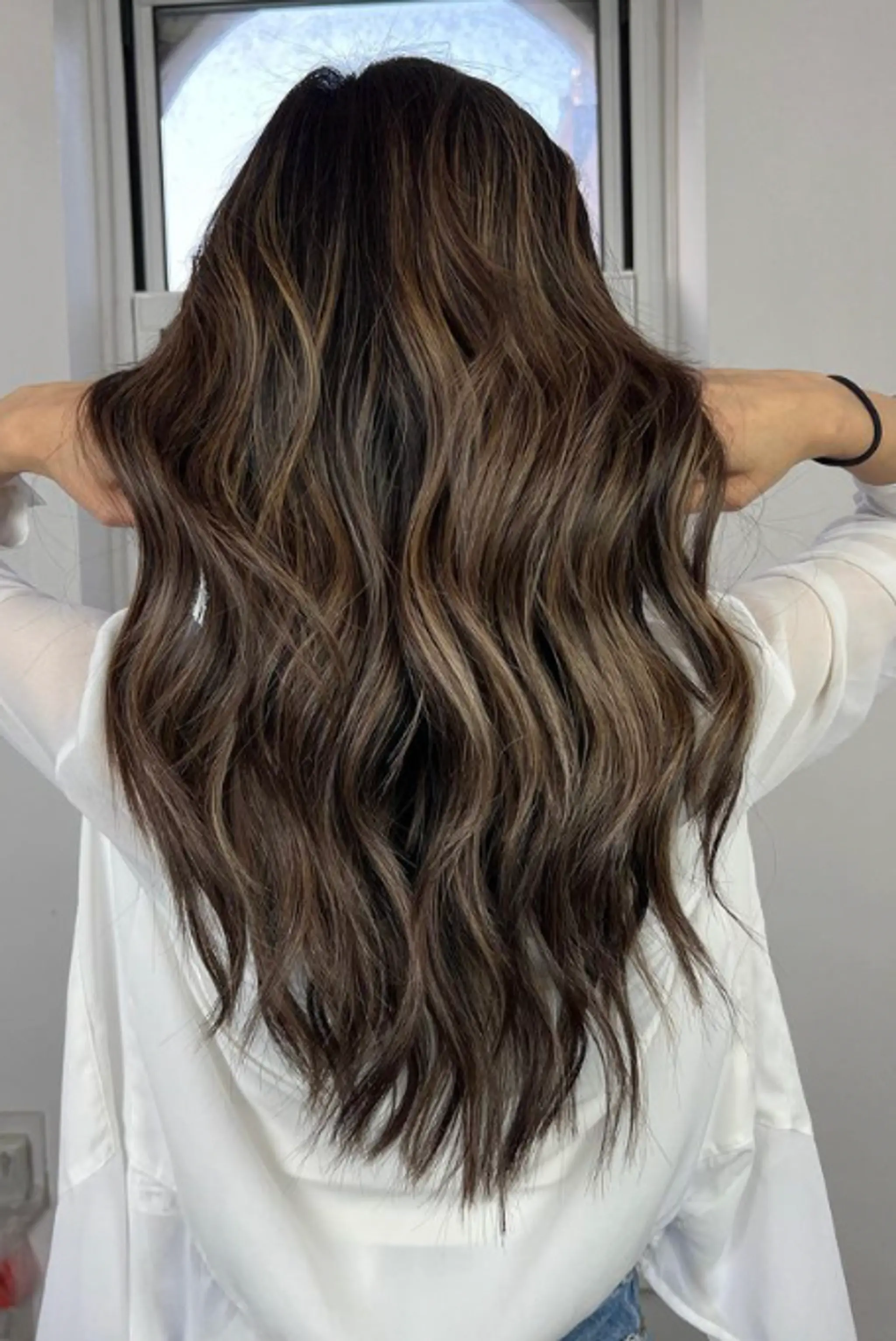 The style features long, cascading waves with a rich brown base color and ash blonde balayage highlights, creating a sun-kissed and multi-tonal effect. The texture is wavy, adding volume and movement to the look.