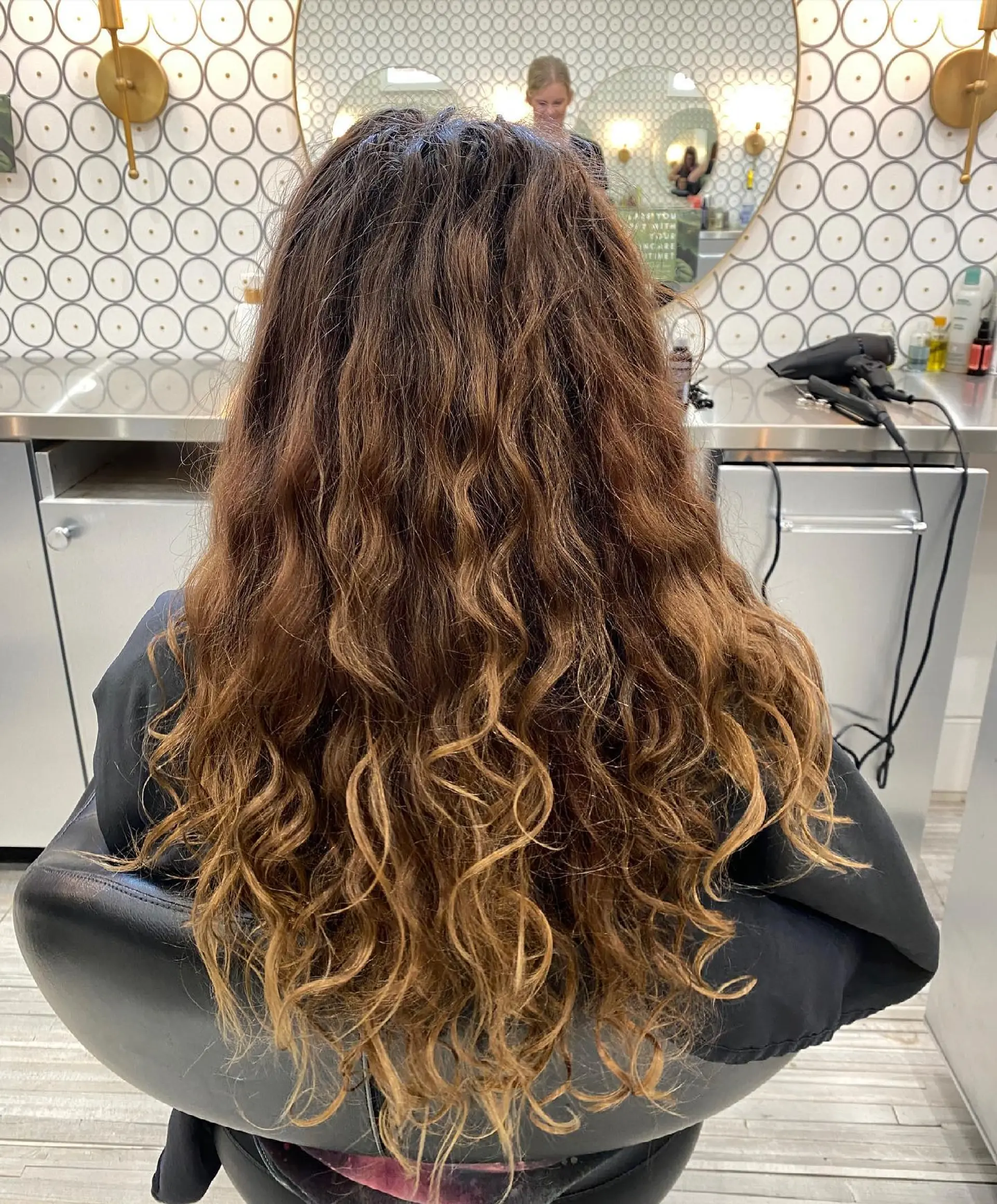 Long curly brunette hair enhanced with warm dimensional balayage highlights throughout the mid-lengths and ends. 1