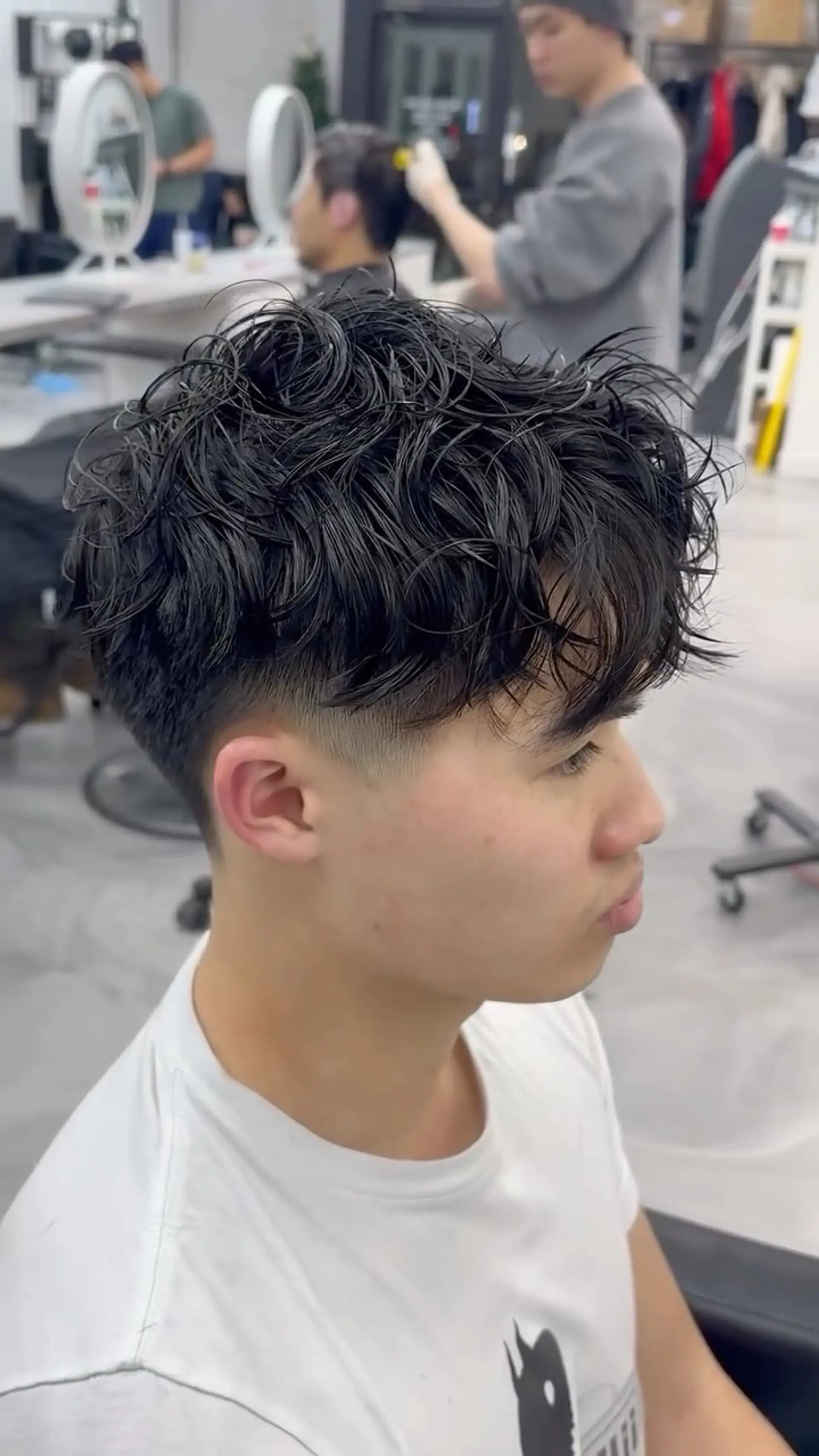 A men's short hairstyle featuring a textured, wavy permed top styled forward with a low taper fade on the sides. 1