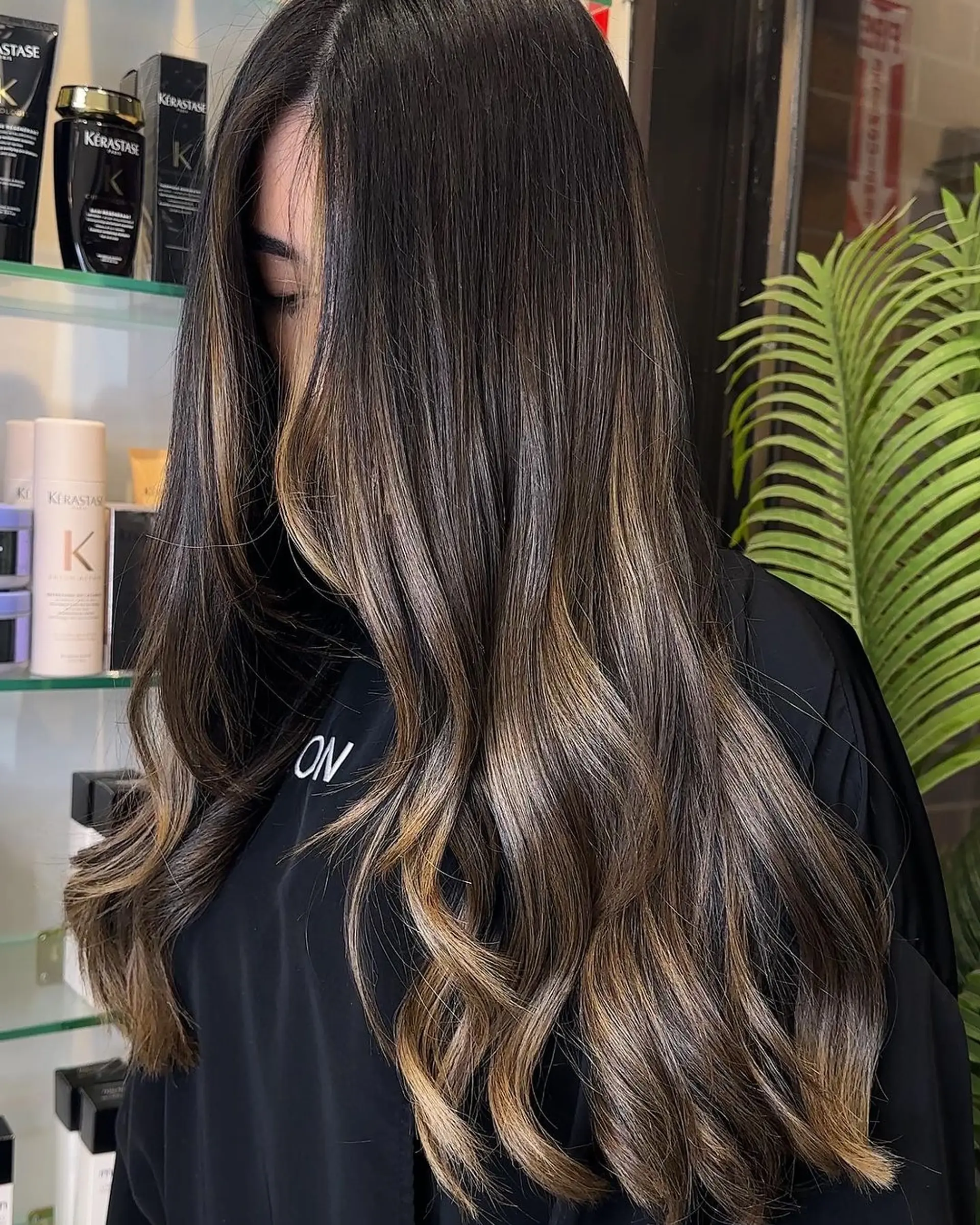 Long, wavy dark brown hair with honey blonde balayage highlights. The waves are soft, and the hair is parted in the middle with subtle layers for added volume. 2