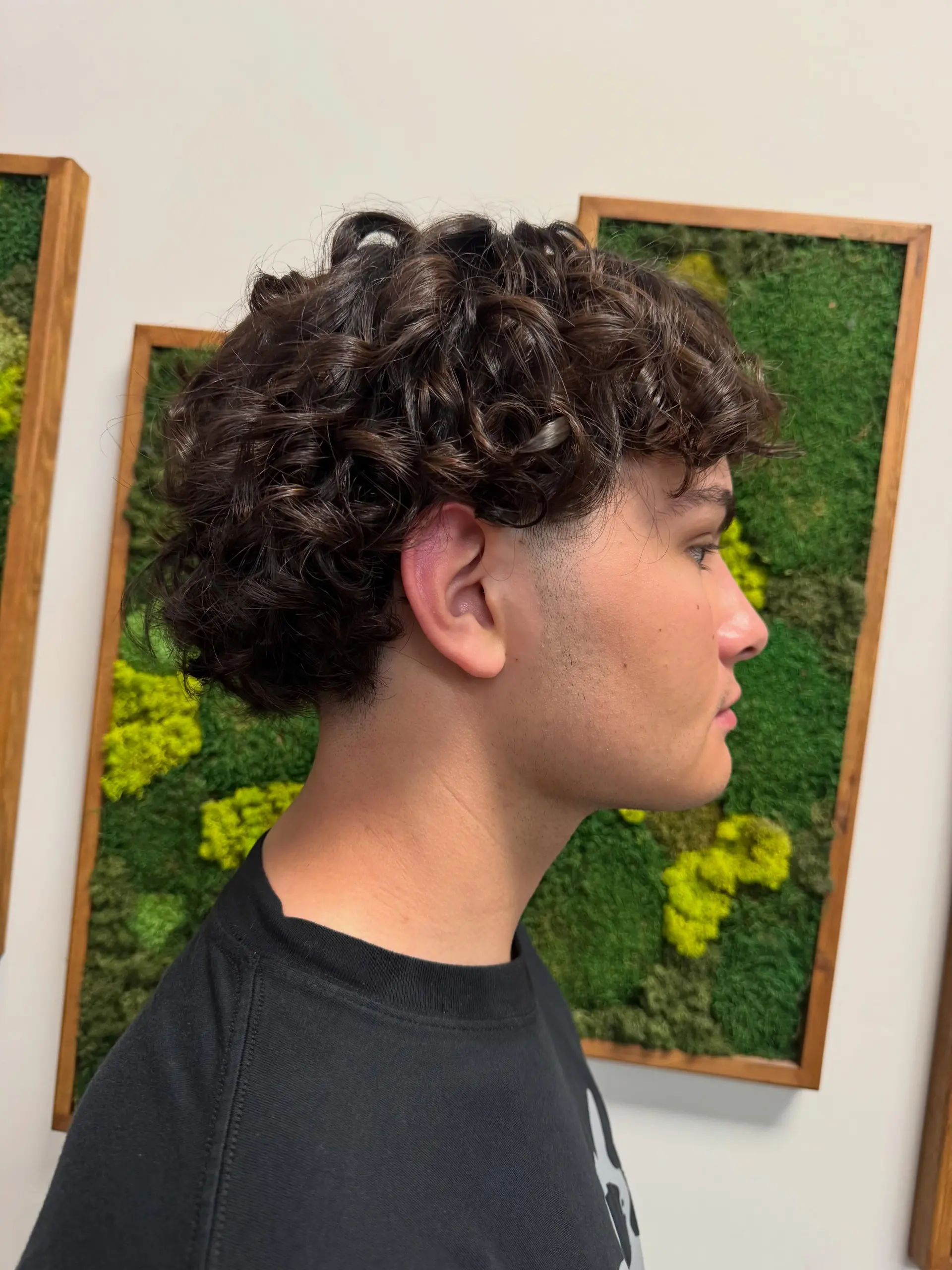 A modern men's perm featuring defined, glossy curls with a clean taper around the ear and neckline. 1