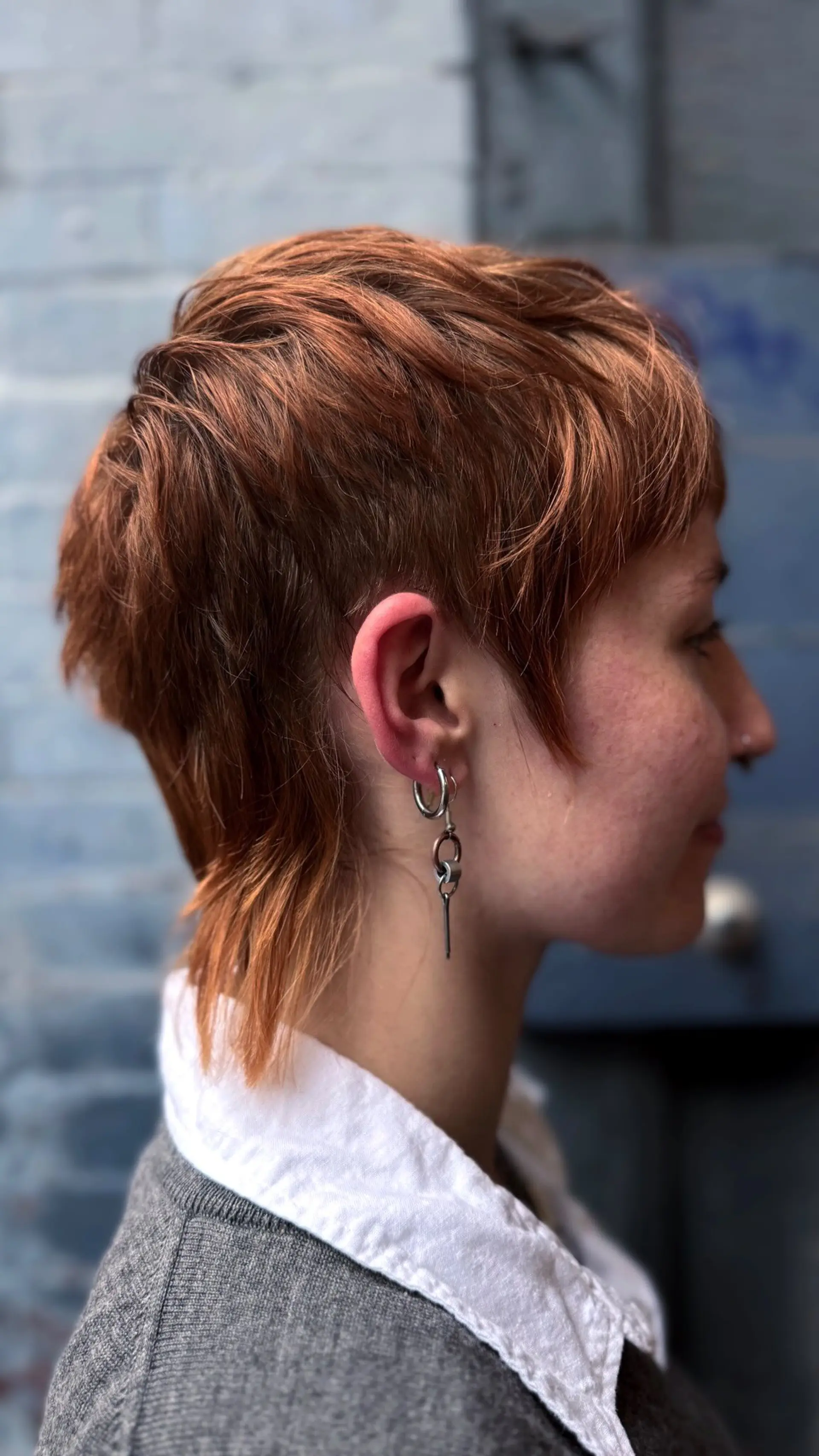 A textured mixie (pixie-mullet hybrid) haircut featuring choppy micro bangs, tapered undercut sides around the ear, and feathered length down the nape. 1