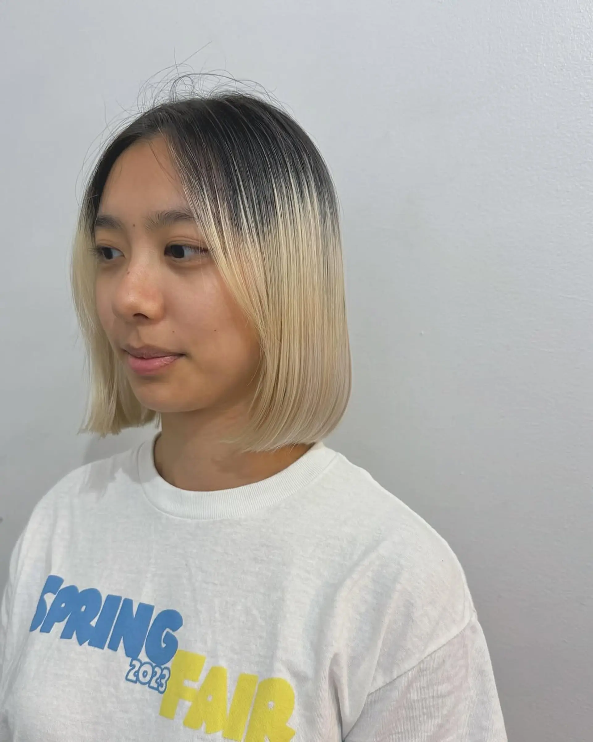 A sleek, blunt bob haircut styled straight with sharp, clean lines ending just below the chin. The style is characterized by a dramatic color contrast, featuring dark natural roots that seamlessly blend into a vibrant platinum blonde. 1