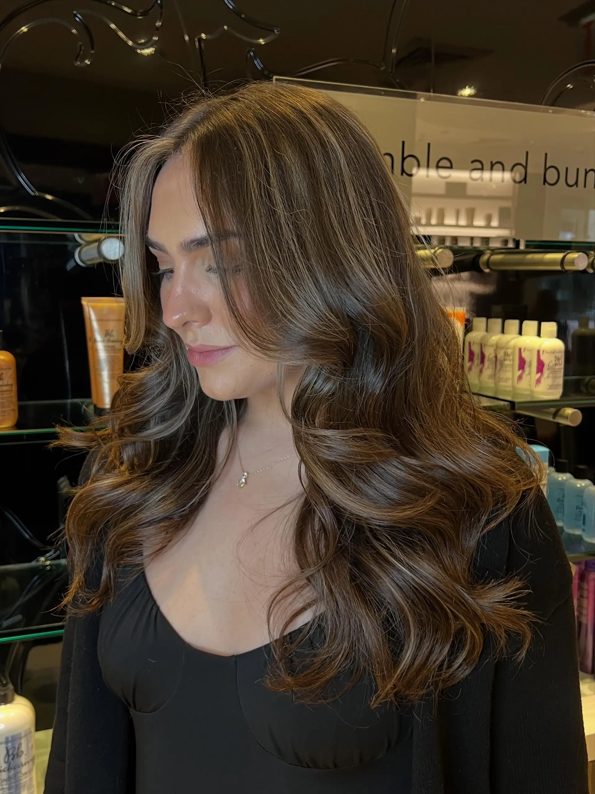 Long dimensional brunette hair with soft balayage highlights styled in bouncy, face-framing waves. 1
