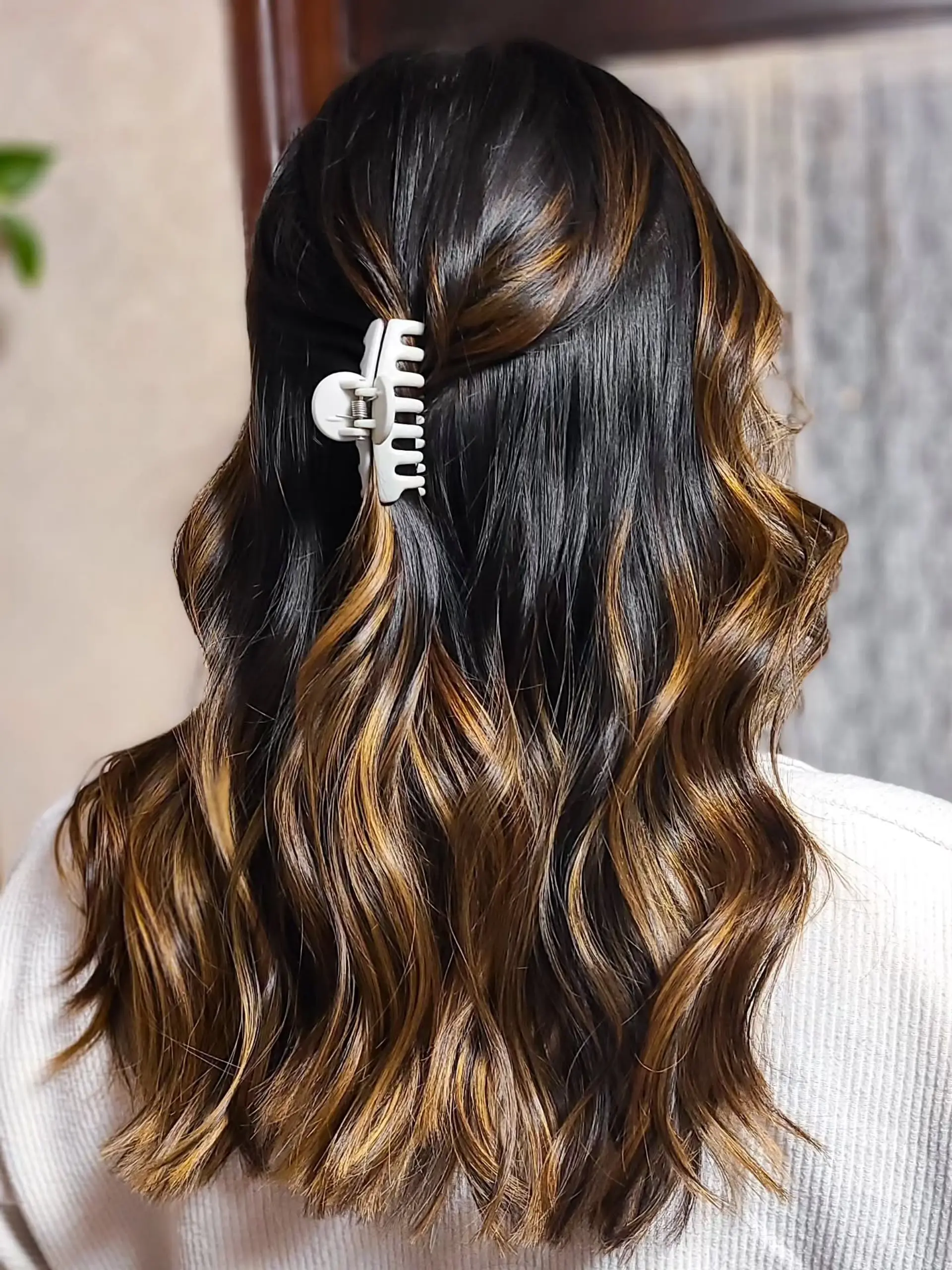 Medium-length wavy hair featuring a dark brown base with caramel and golden brown balayage highlights, styled with a half-up claw clip for an effortless look. 1