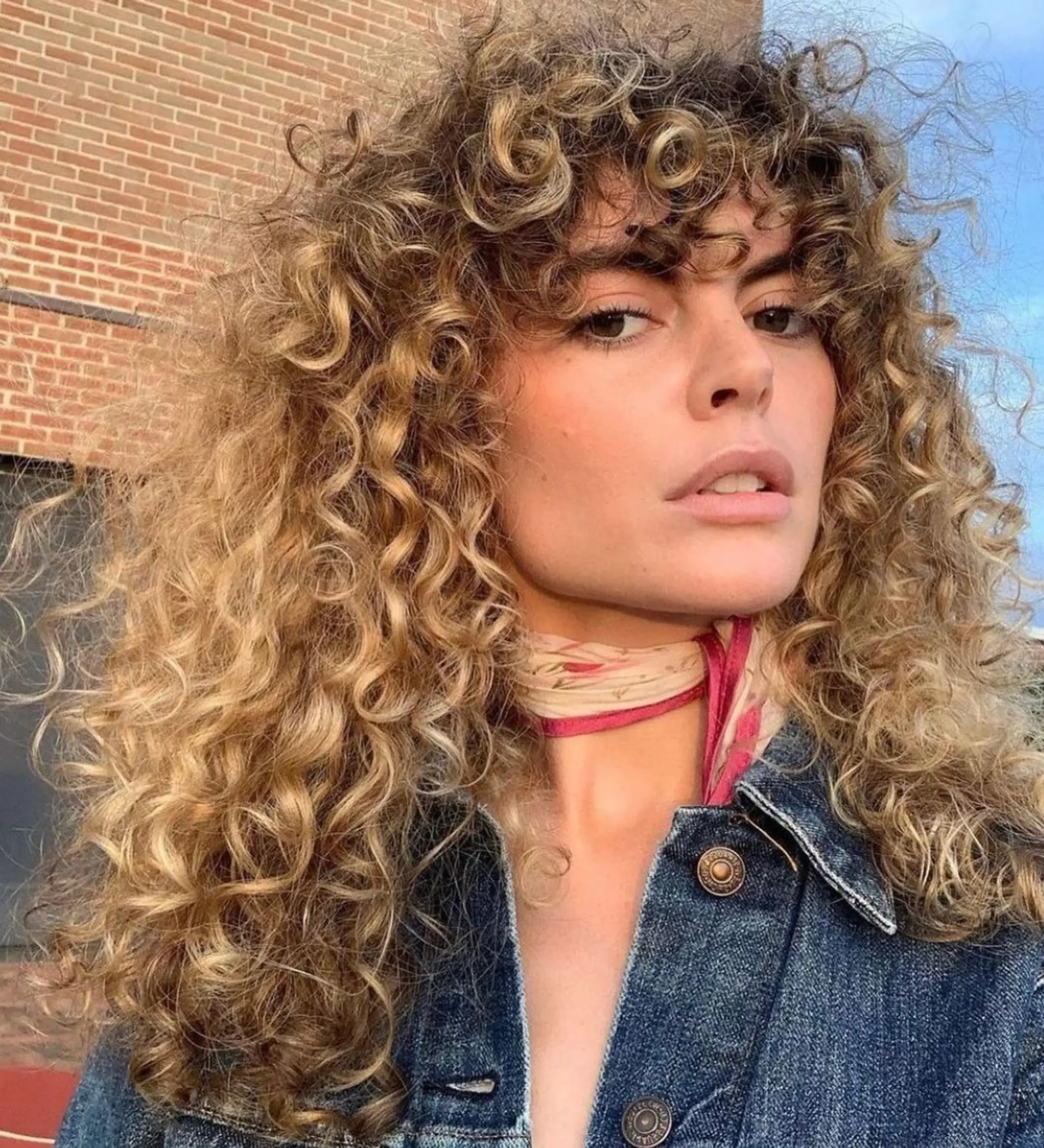 Voluminous long curly hair in golden blonde and honey blonde shades, featuring defined curls and fringe bangs.
