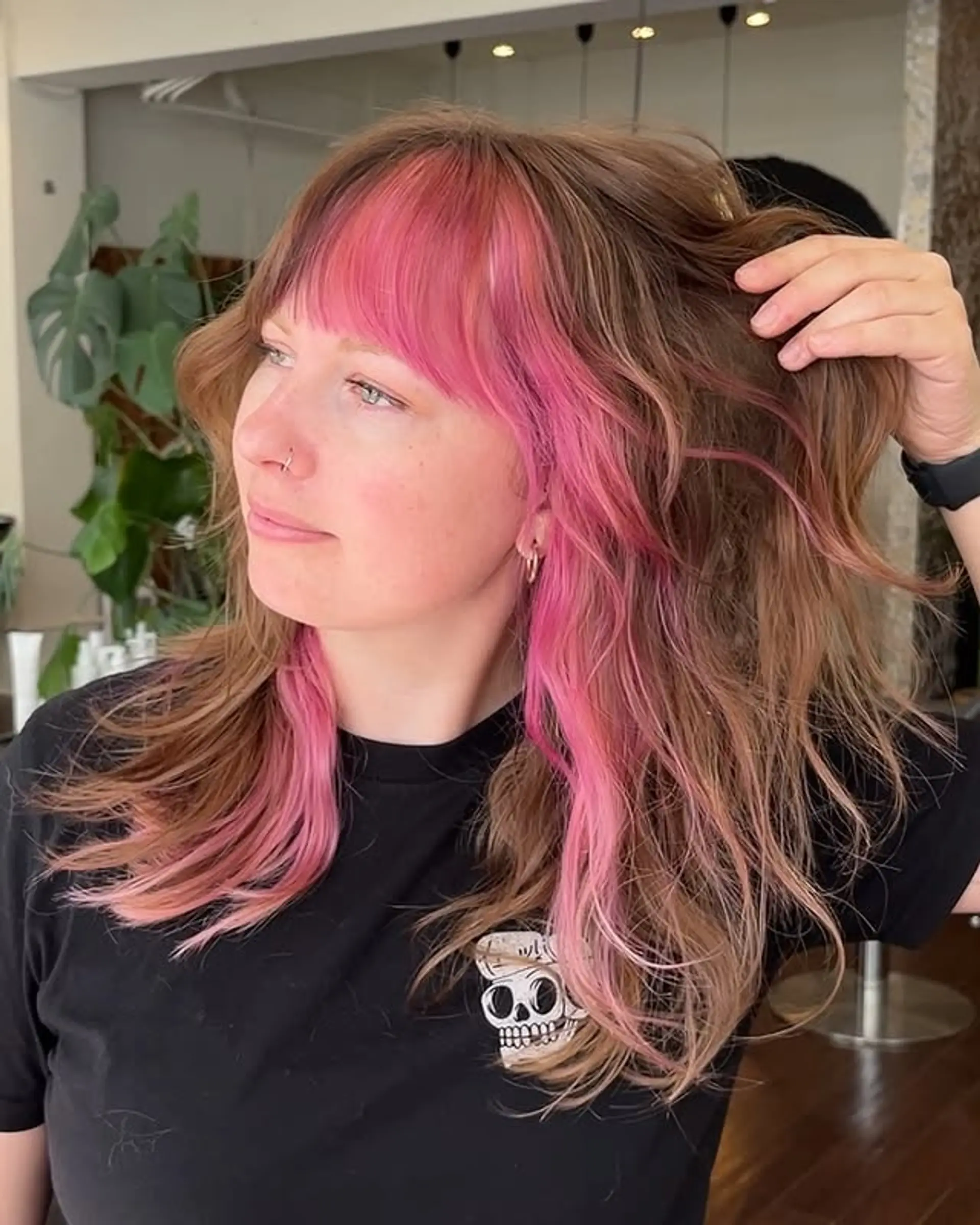 This hairstyle features medium-length wavy hair with full curtain bangs and striking pink money piece highlights to frame the face. 1
