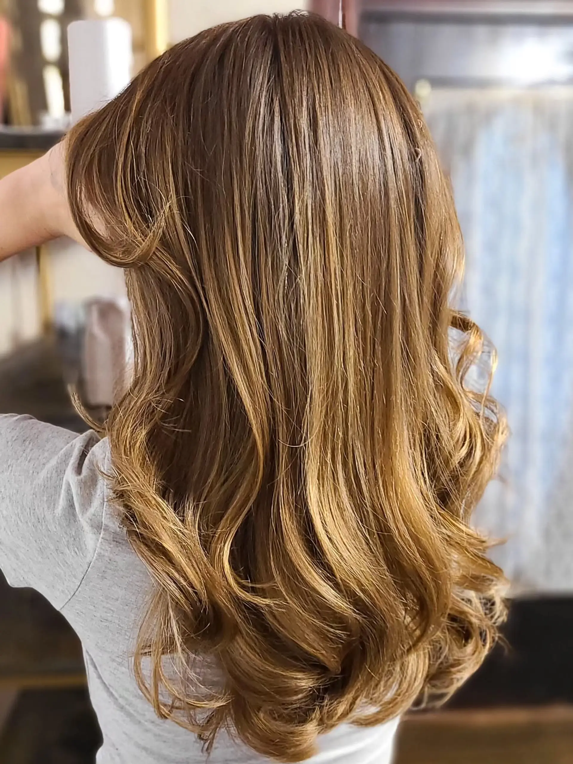 Long, layered hair styled with soft, voluminous waves, showcasing a dimensional bronde balayage that blends brown with golden blonde and caramel highlights. 3