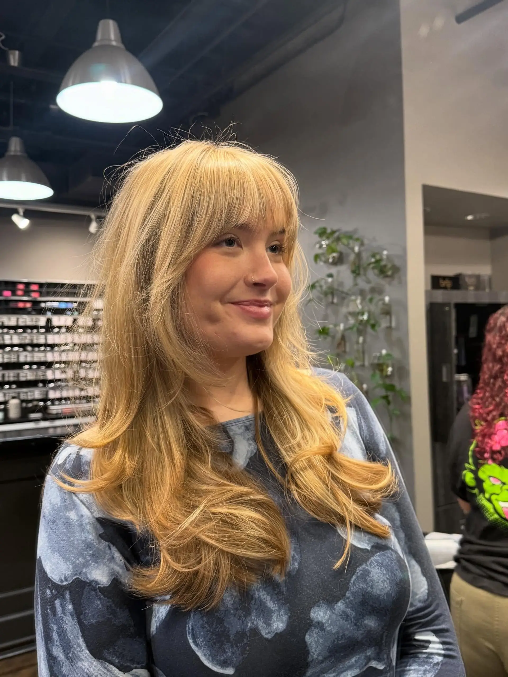 Long warm blonde hair cut with soft face-framing layers and full straight-across bangs, styled with voluminous turned-in ends. 1