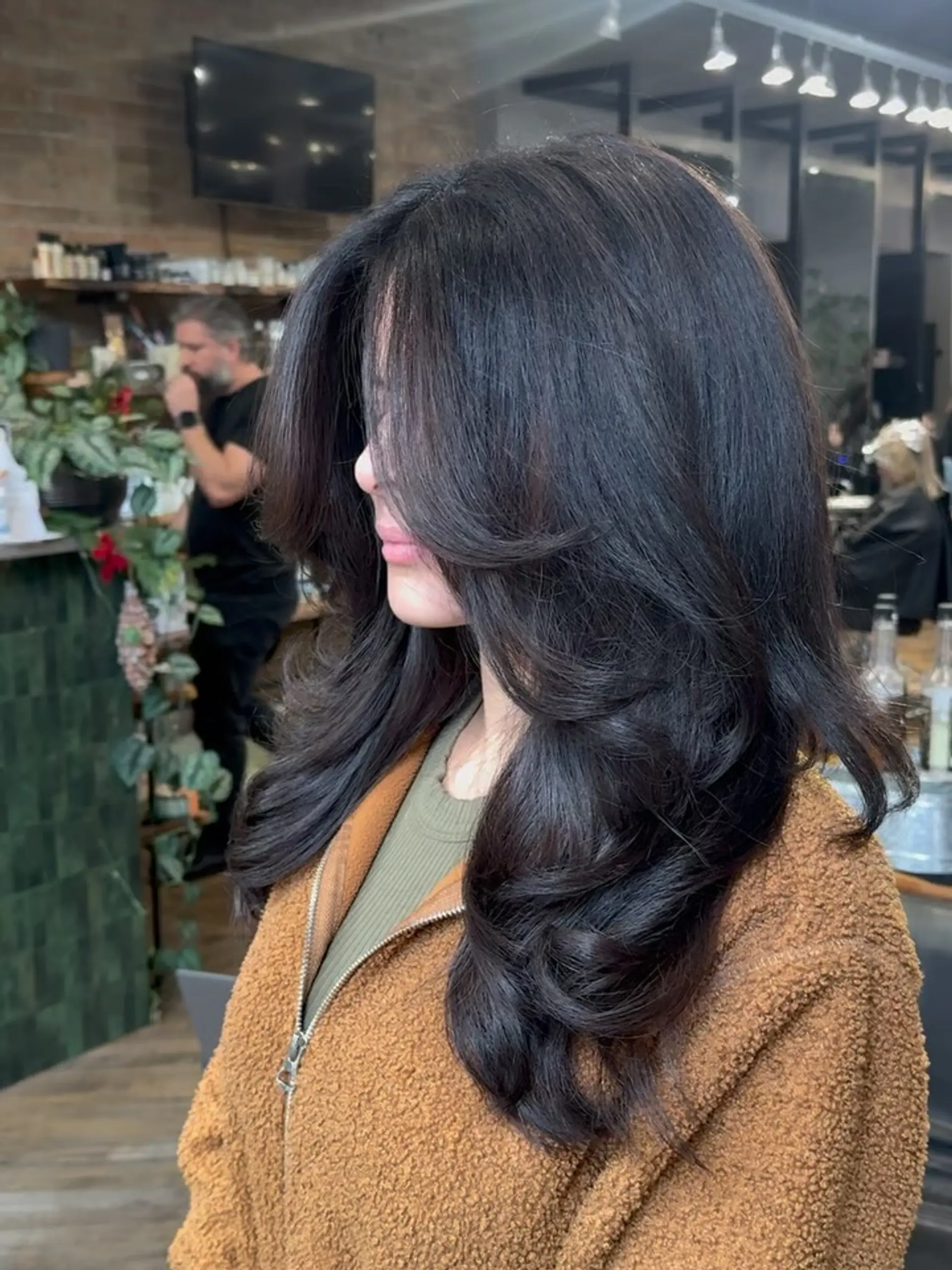 A lush, long dark brunette haircut styled with sweeping curtain bangs, seamless face-framing layers, and soft, bouncy waves. 1