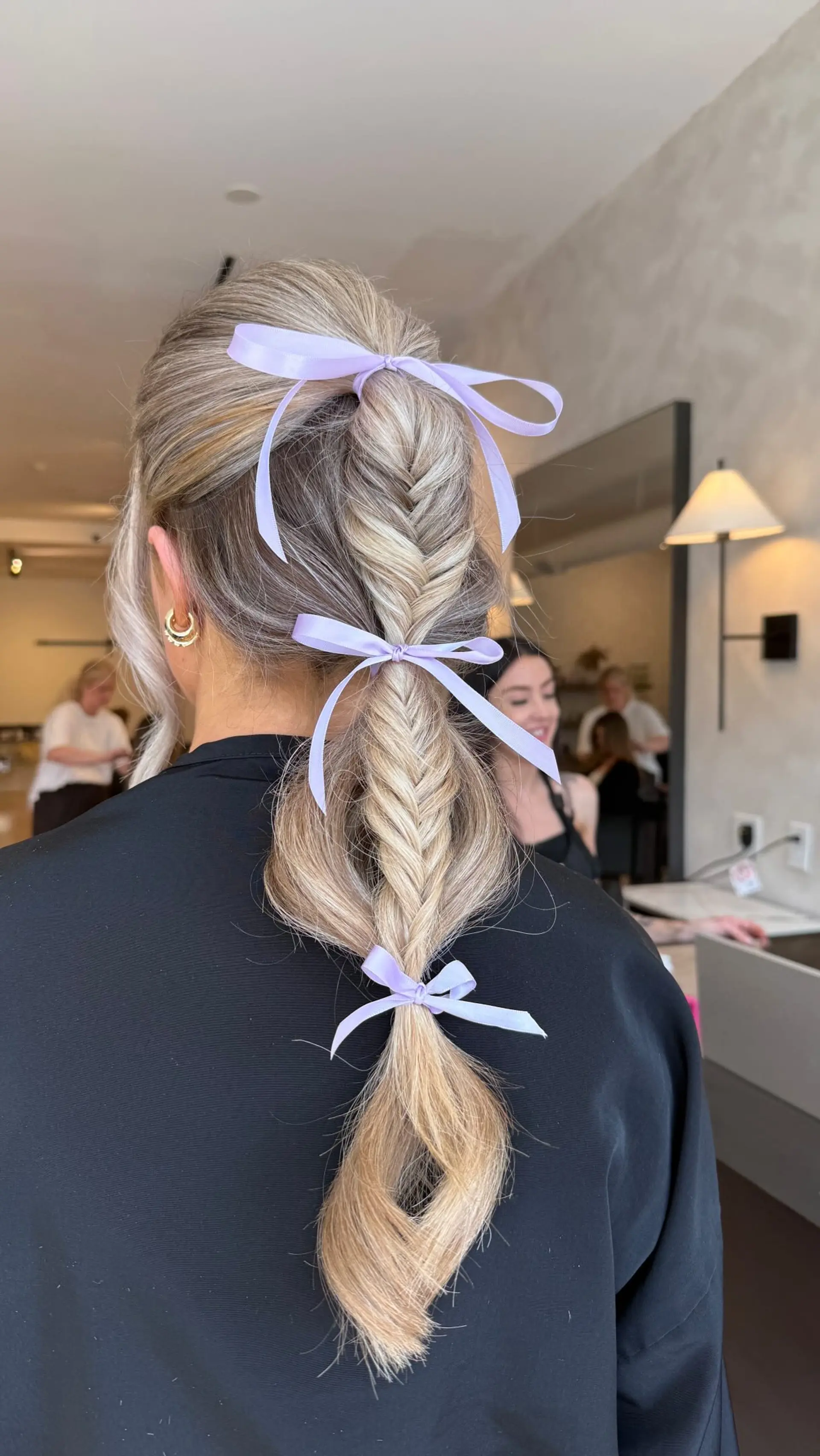 A dimensional blonde fishtail braid styled into a relaxed ponytail accented with soft lavender ribbon bows. 1