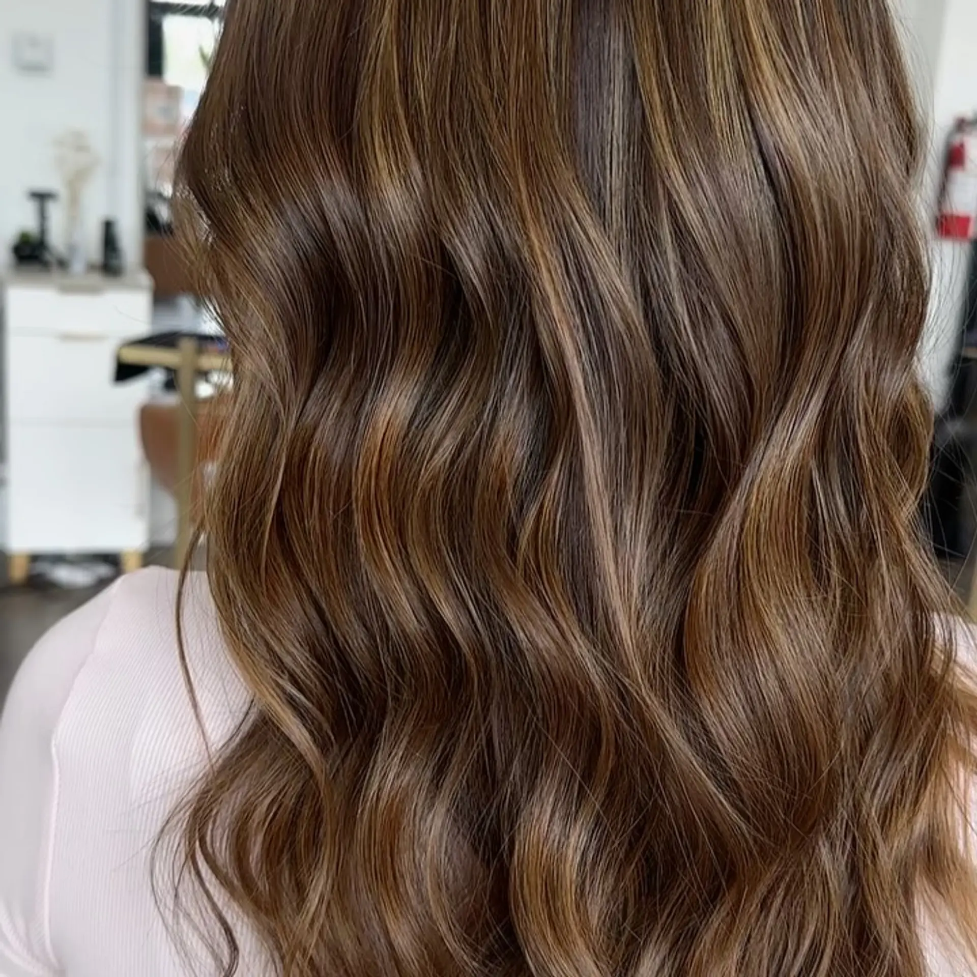 Long, glossy brunette hair styled in loose waves with soft dimensional caramel and light brown highlights. 1