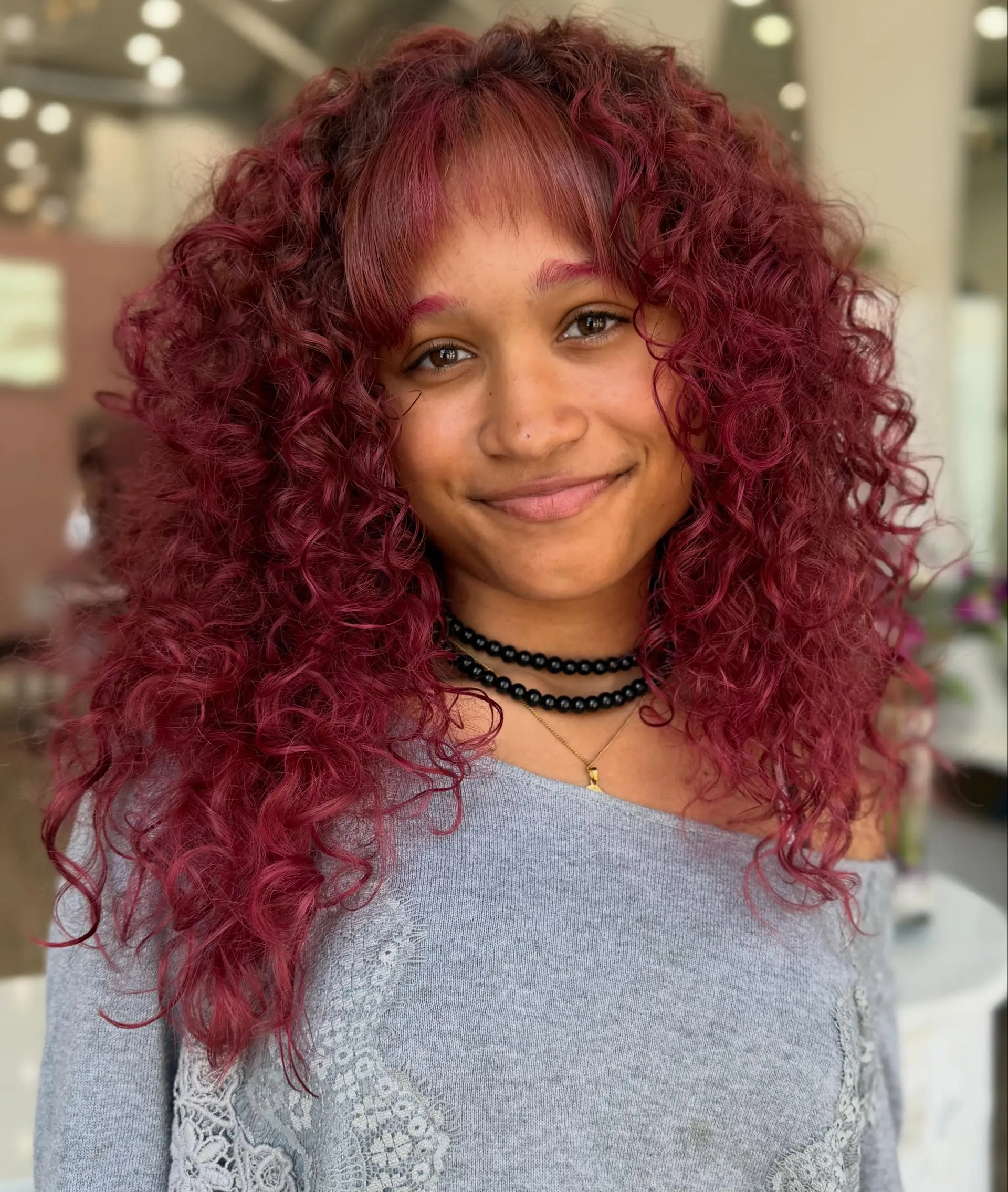 Medium-length, voluminous defined curls in a rich magenta-burgundy shade paired with light wispy bangs. Estimate scope: Haircut only. The vivid burgundy/magenta color described is not priced by the mapped service. Published salon-level starting/range estimate; confirm the chosen provider’s quote. 1