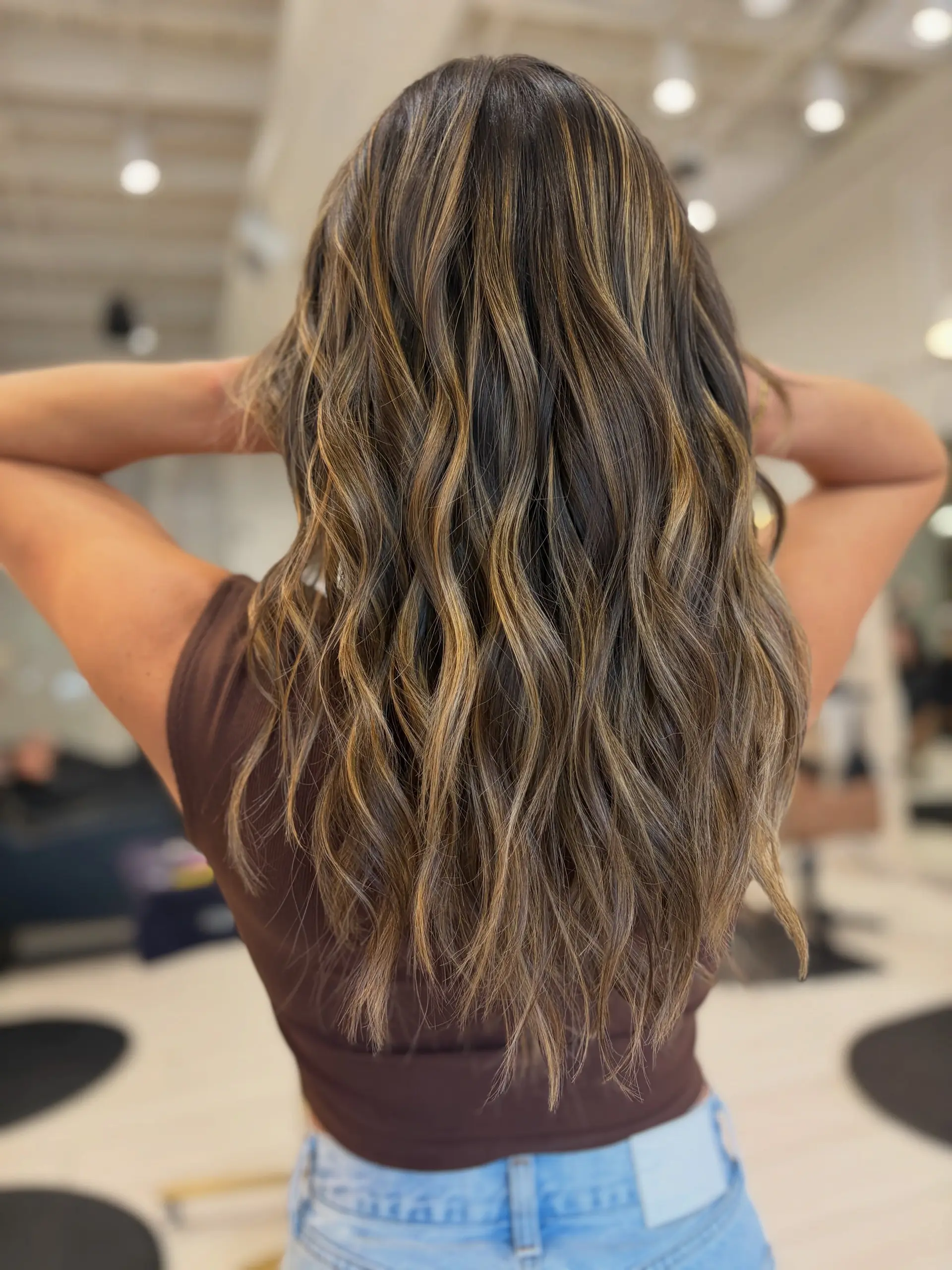 Long dimensional brunette hair styled in soft, loose waves featuring warm caramel and golden highlights throughout a deep brown base. 1
