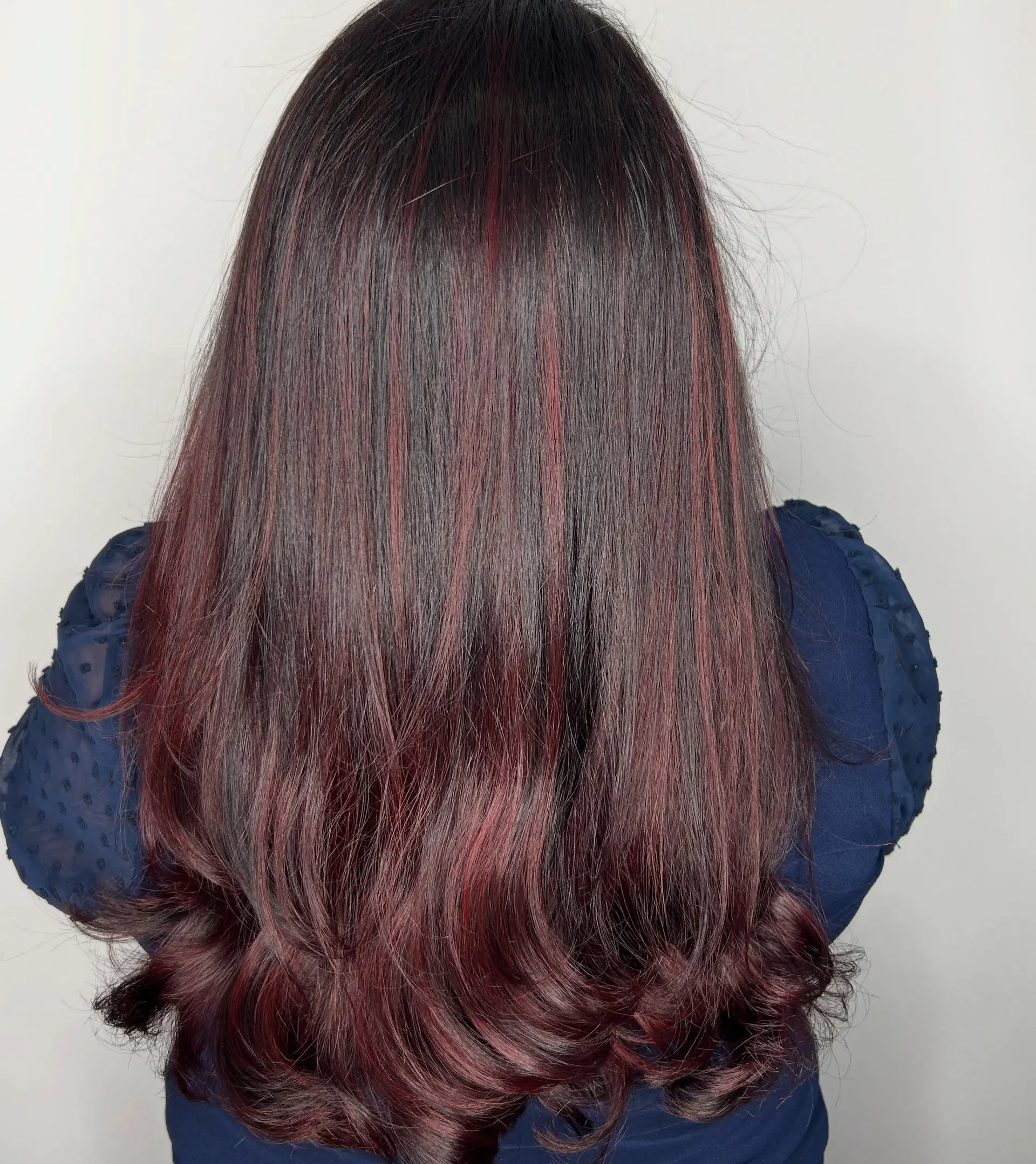 Long dark hair accented with seamless burgundy balayage highlights, styled smooth with soft curls at the ends. 1