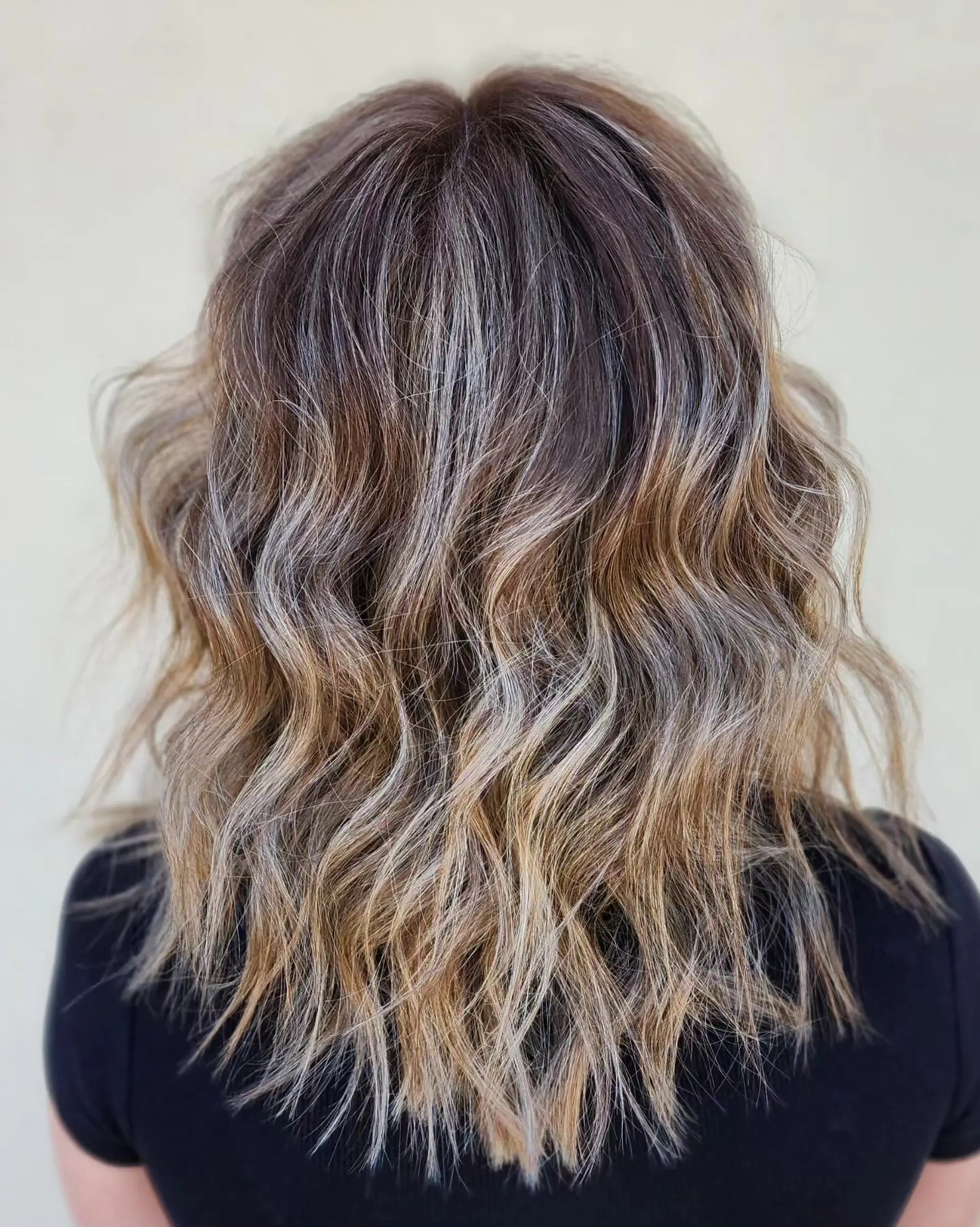 Medium-length textured haircut styled in loose beach waves with a multi-tonal ash blonde foilayage on a dark brown base. 1