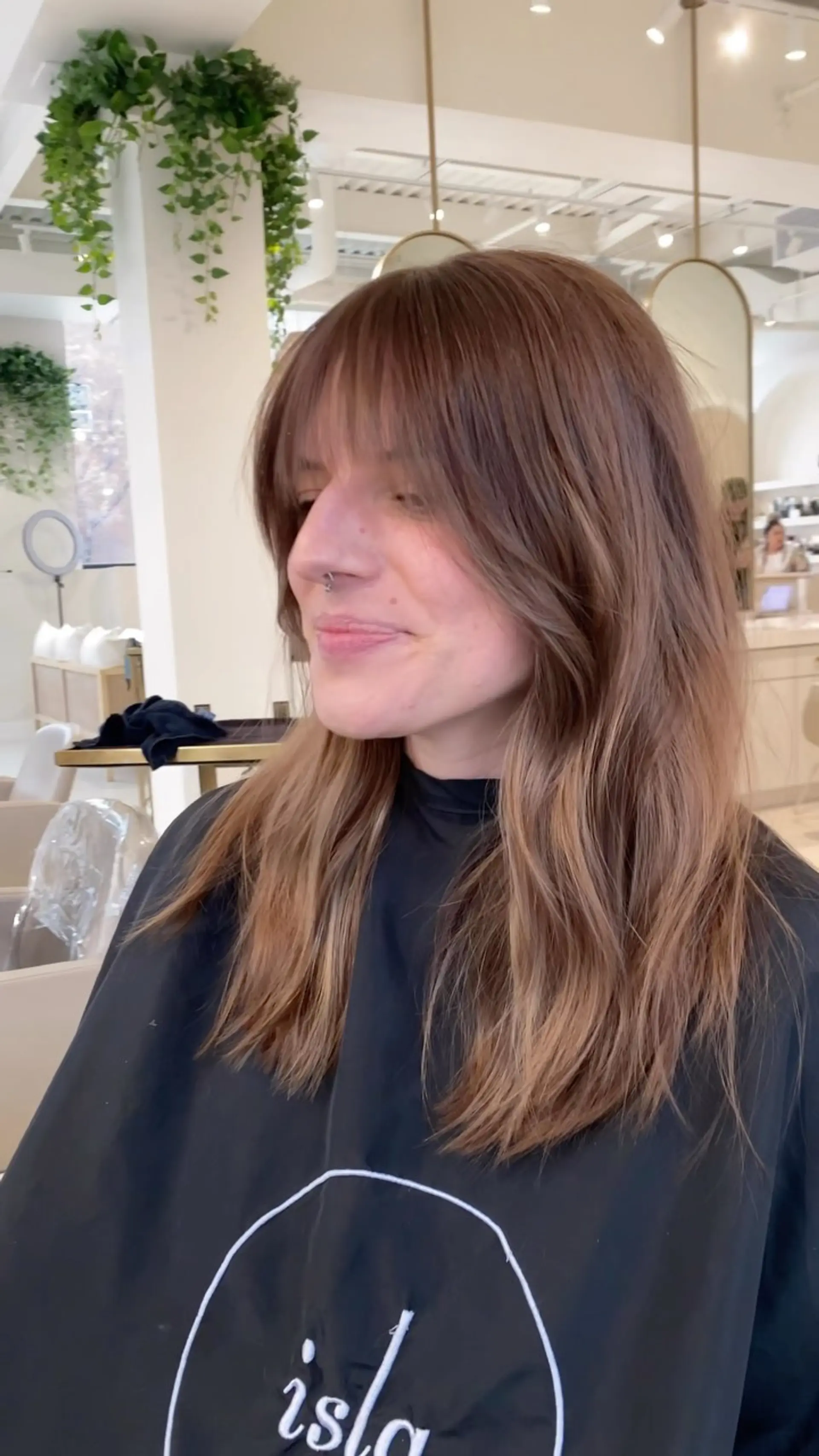 Medium-length warm brunette hair styled with soft, face-framing layers and wispy eyebrow-skimming bangs. 1