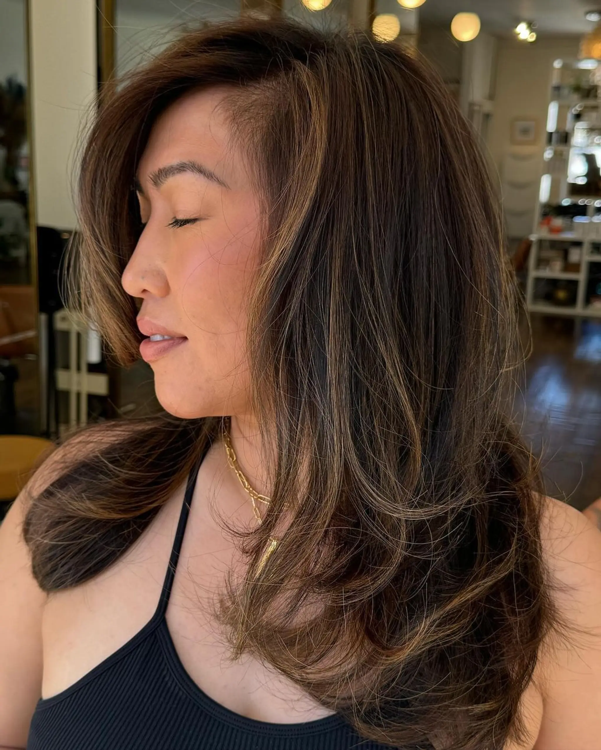 A long, layered haircut with a side part and golden highlights, offering natural volume and face-framing elegance in straight textures.