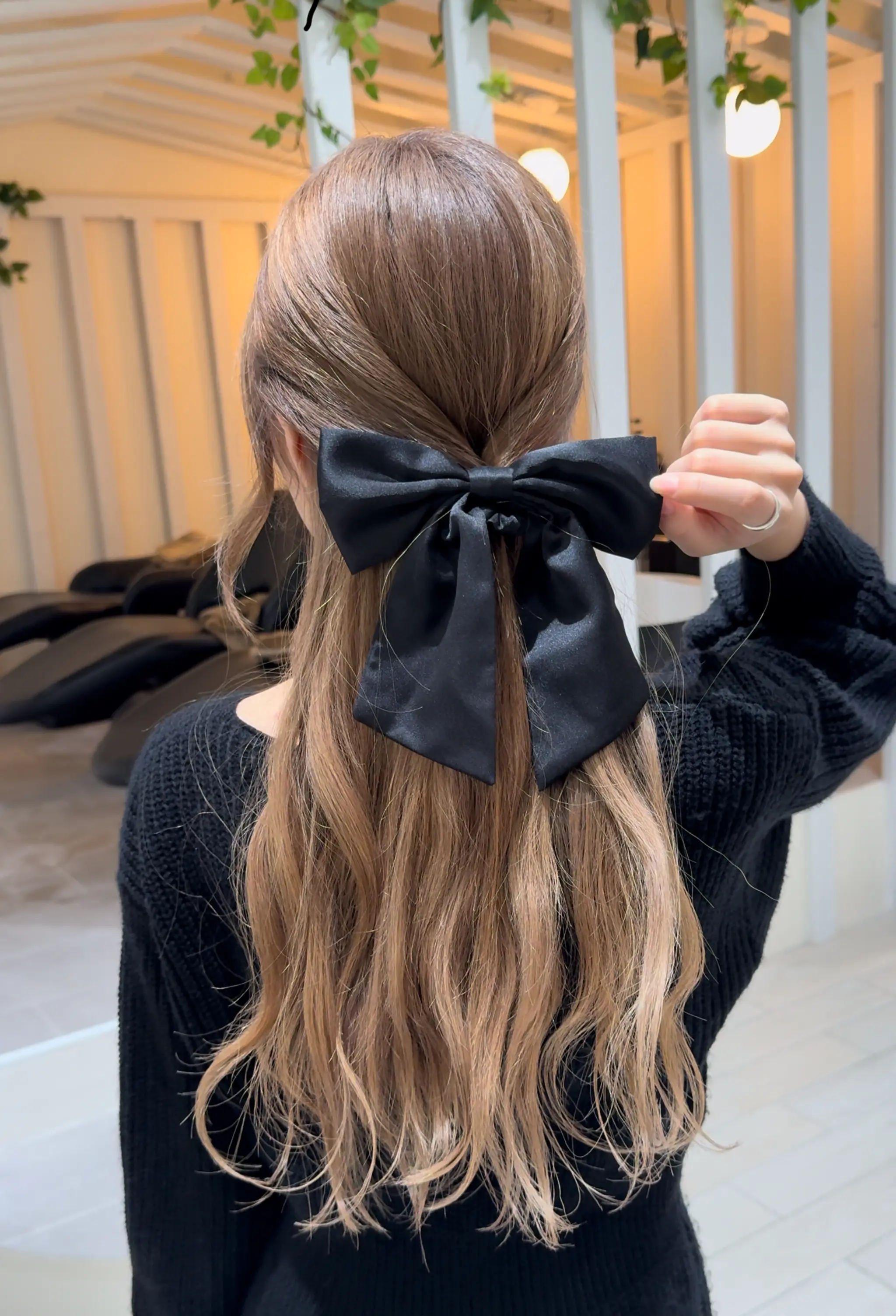 Long, wavy hair with light brown base color and blonde highlights, styled in a half-up fashion with a satin black bow at the back.