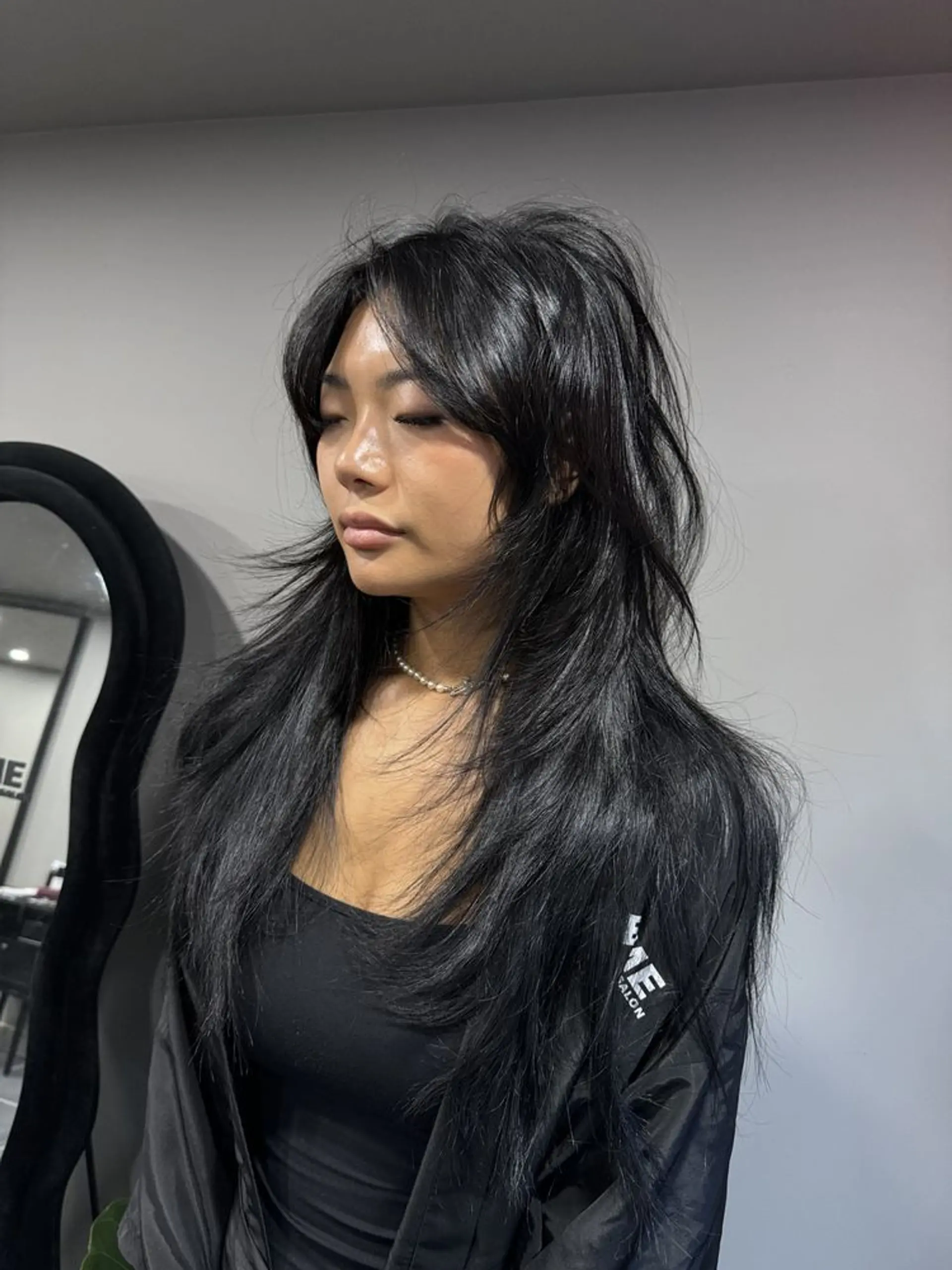 This long, dark black hairstyle features voluminous, textured layers that cascade down past the shoulders. The hair has a wavy texture, enhancing the overall fullness and movement. The style is characterized by feathery ends and side-swept bangs that frame the face beautifully. 1