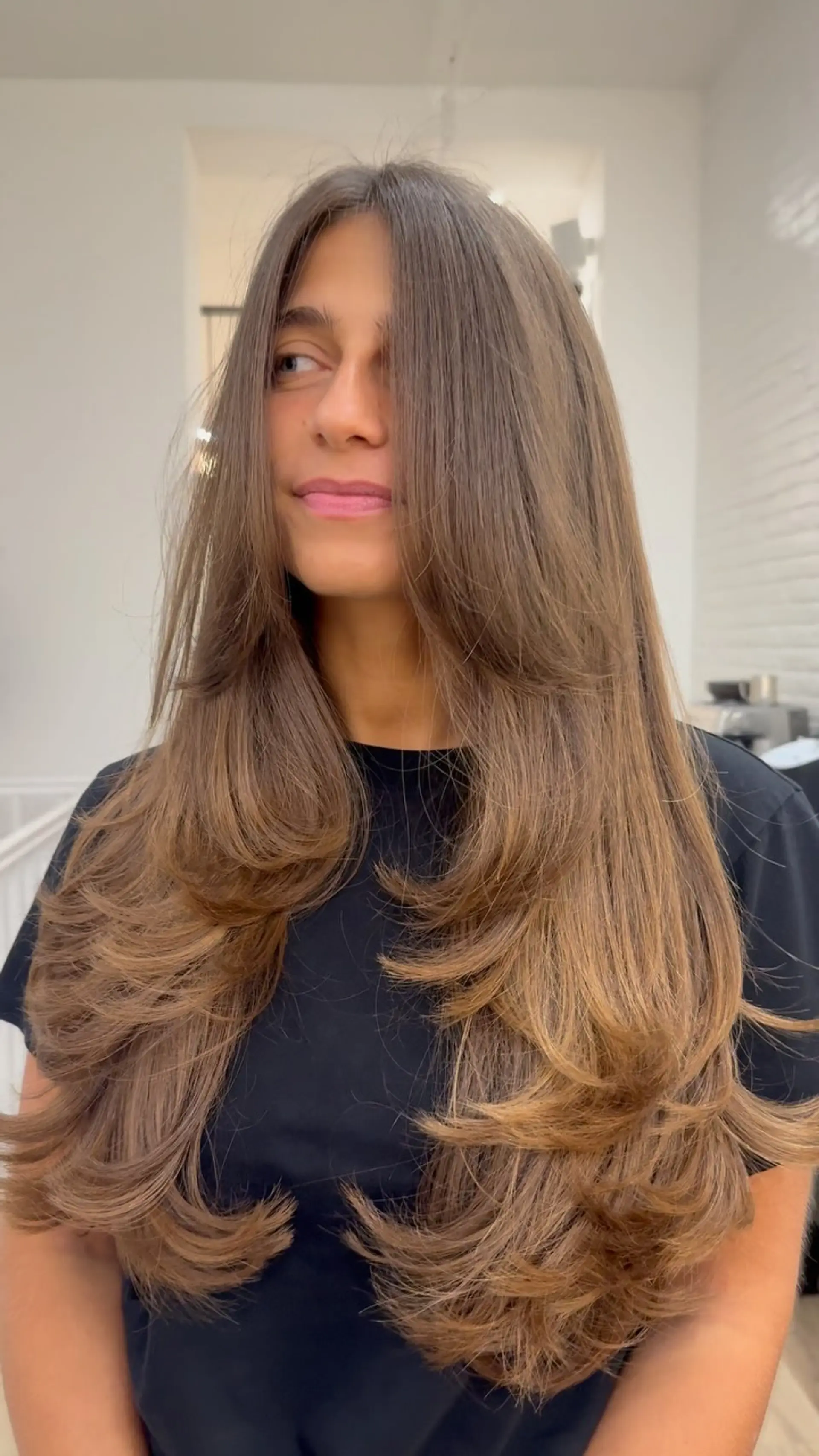 Long brunette hair cut in cascading, bouncy layers with face-framing curtain pieces, styled in a polished, voluminous blowout. 1