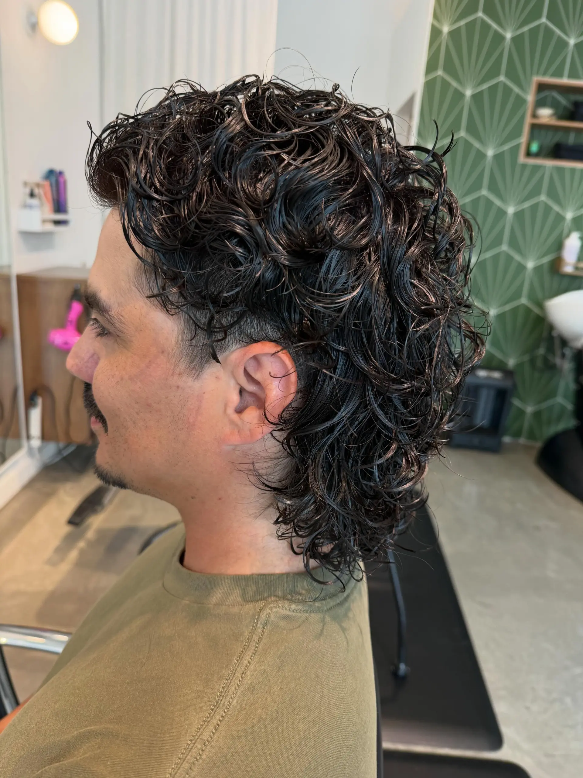 A textured, modern mullet featuring wet-styled, defined curls created through a fresh perm, tapered around the ears with length extending at the nape. 1