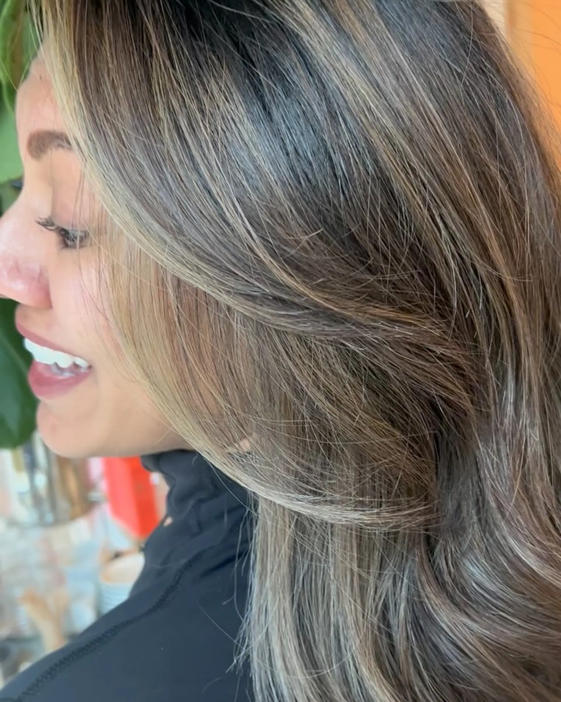 A close-up side view of long, layered hair with cool-toned bronde highlights and face-framing layers styled in a soft blowout. 1