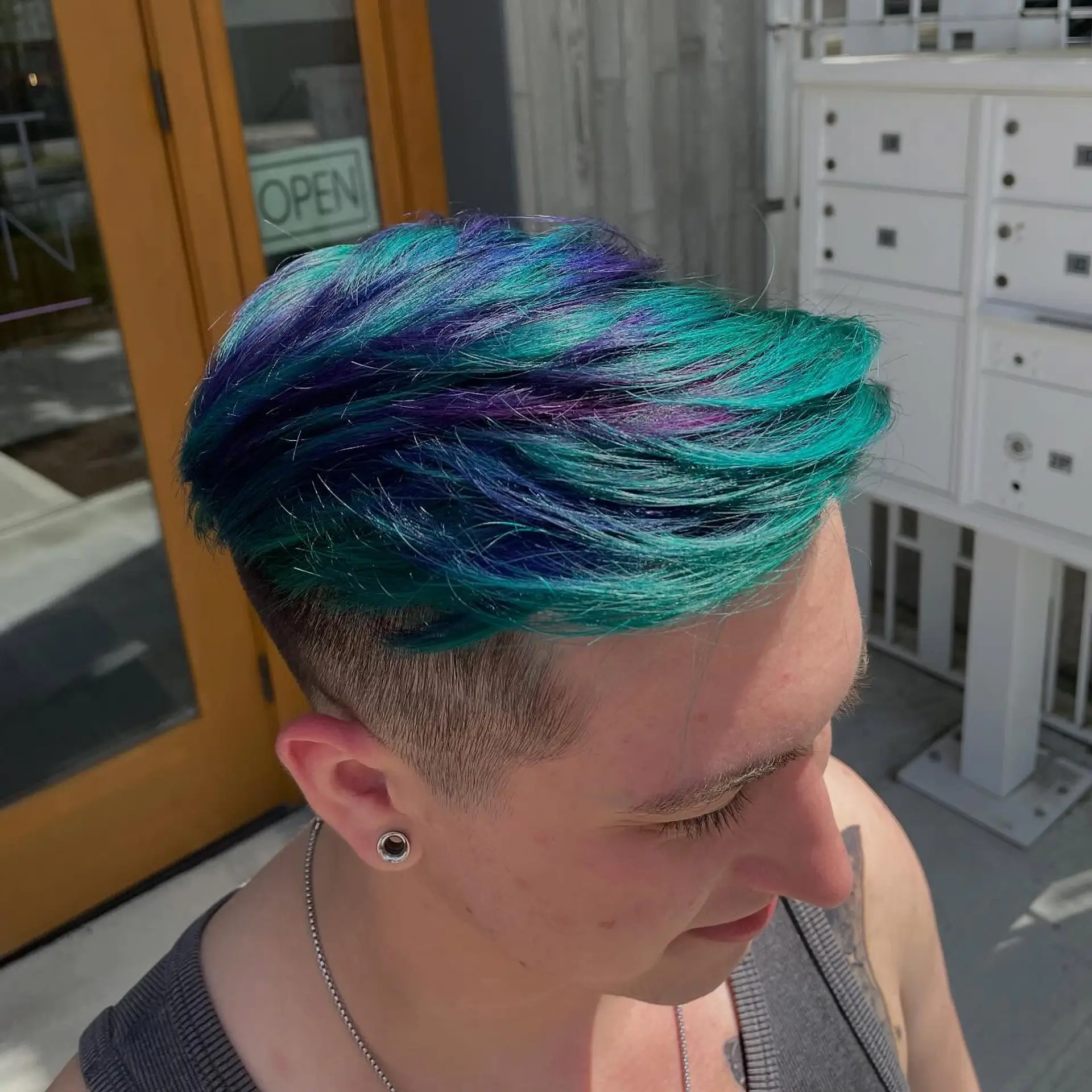 A short textured hair crop featuring a side undercut and vibrant multi-tonal peacock teal and purple top layers. 1