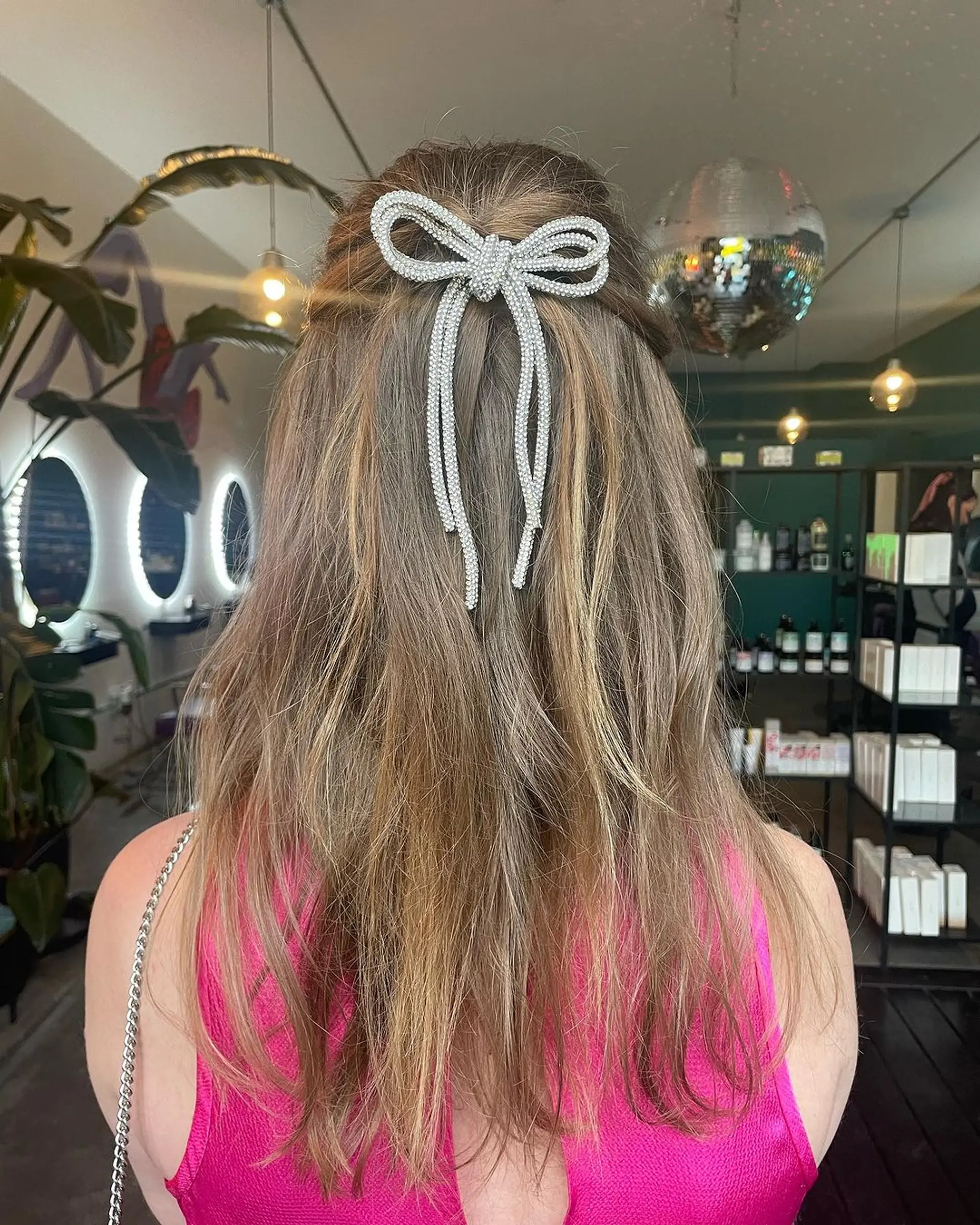 Medium-length wavy hair styled in a half-up fashion with a distinctive silver bow accessory, blending dark blonde, light brown, and ash shades for a casual yet refined look. 1