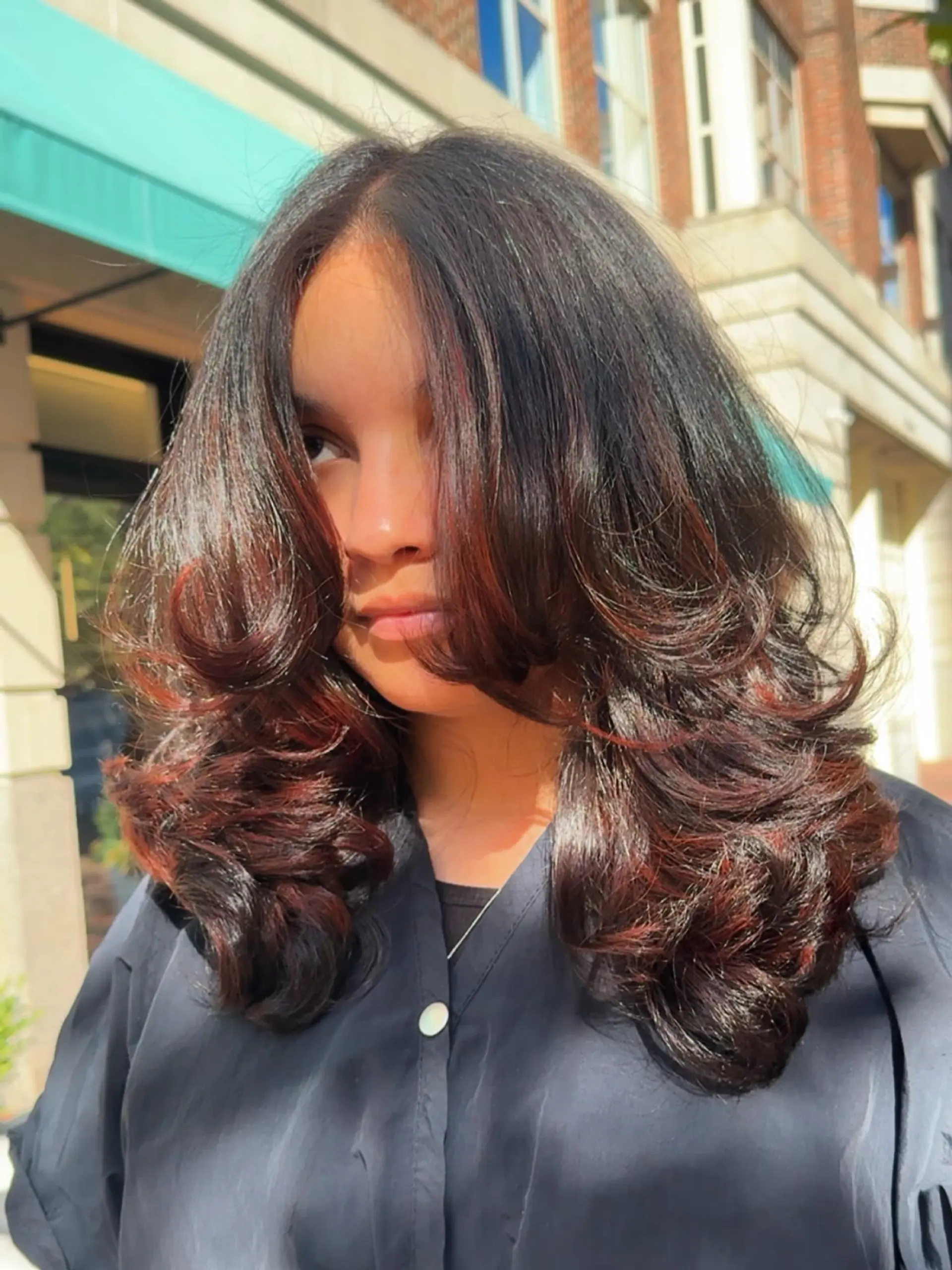 Medium-length dark layered hair styled in a bouncy, voluminous blowout with warm auburn and red foilayage highlights. 1