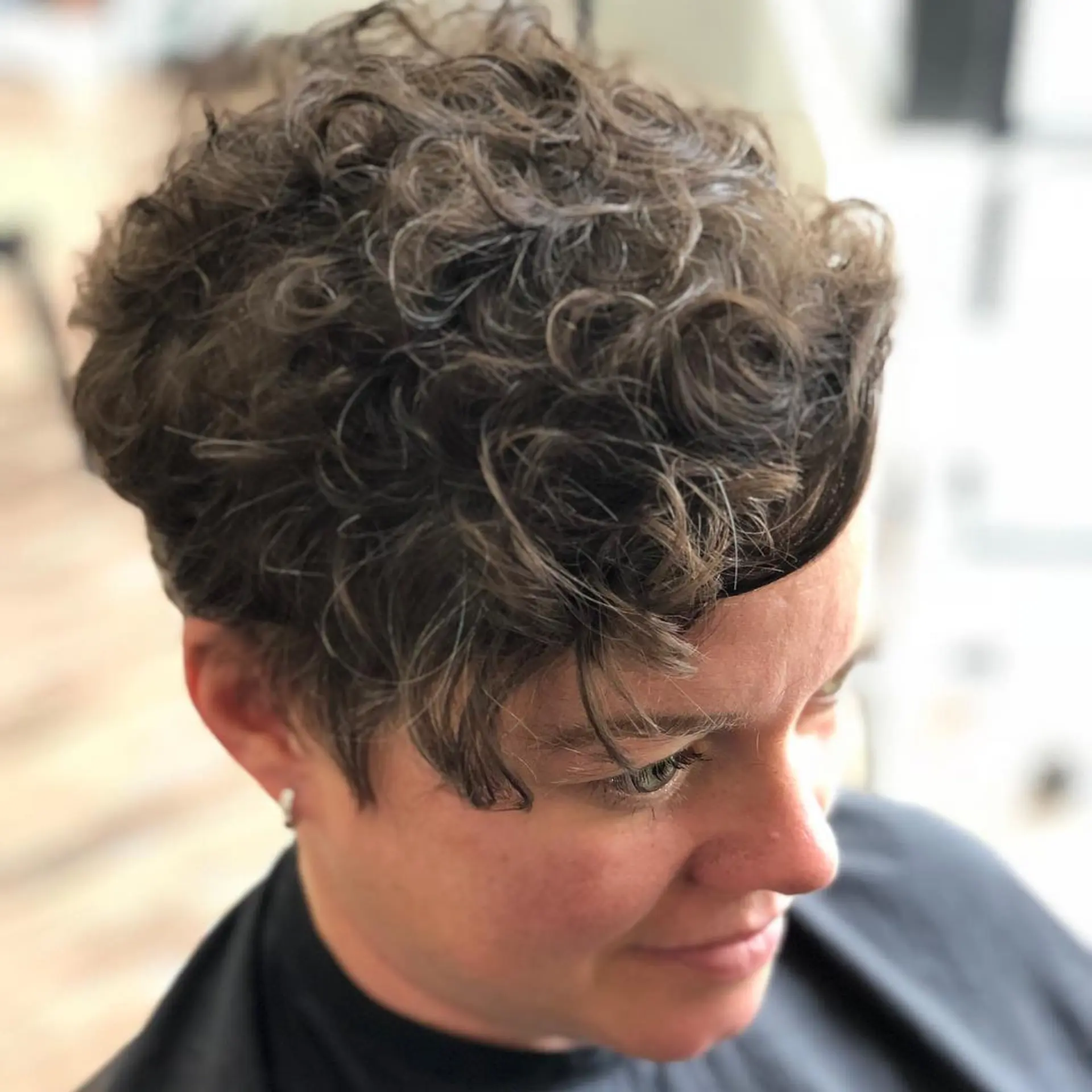 A textured, short curly pixie haircut styled with a longer asymmetrical fringe sweeping across the forehead. 1