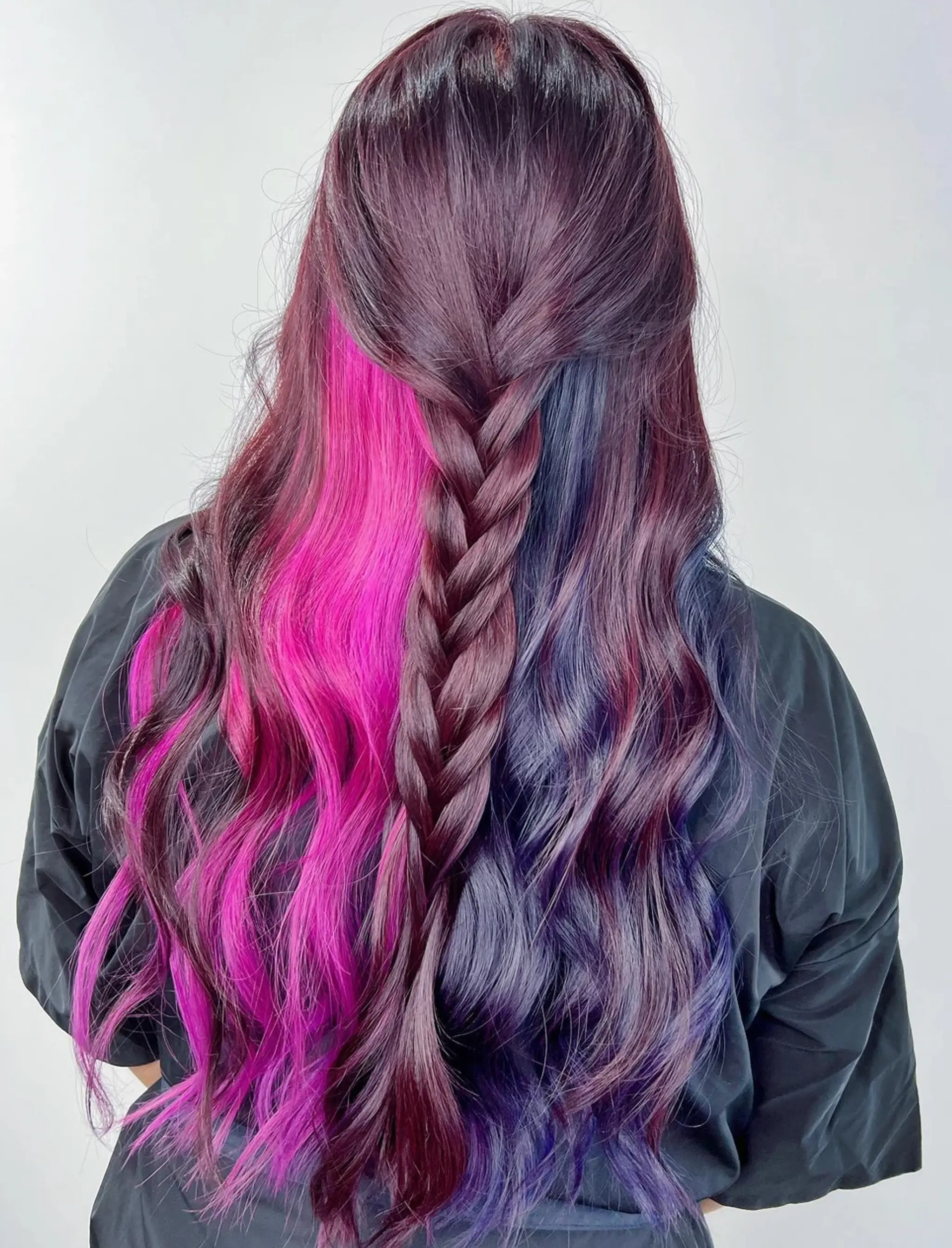 A long, wavy hairstyle with an ombre blend of purple, pink, and blue colors. It includes a central braid in a half-up, half-down style, adding a touch of elegance to the colorful waves.
