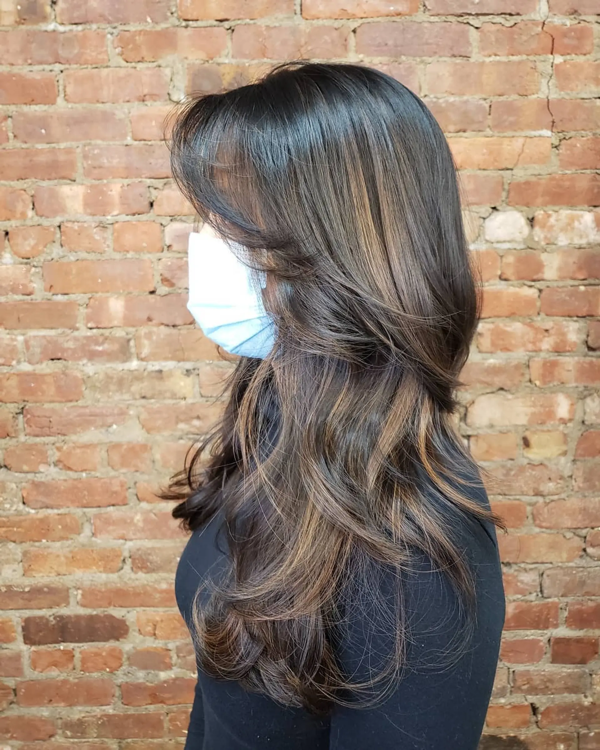 Long dark brown hair featuring feathered, face-framing layers and soft subtle balayage highlights in warm caramel tones, finished with voluminous flipped ends. 1