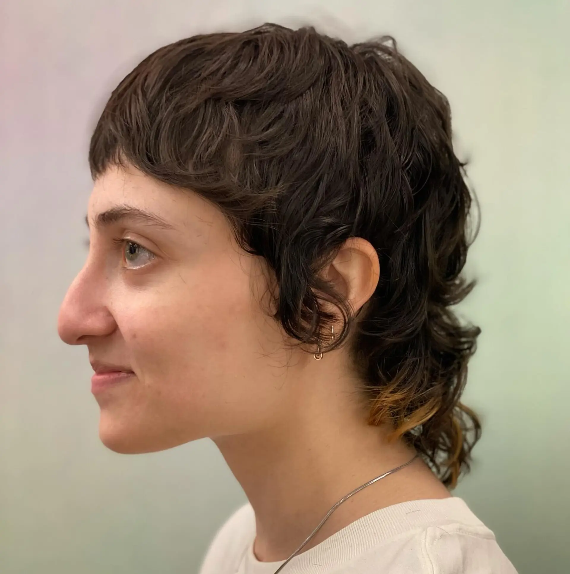 A textured, wavy shaggy mullet cut featuring choppy micro bangs, soft face-framing curls around the ears, and layered length at the nape. 1