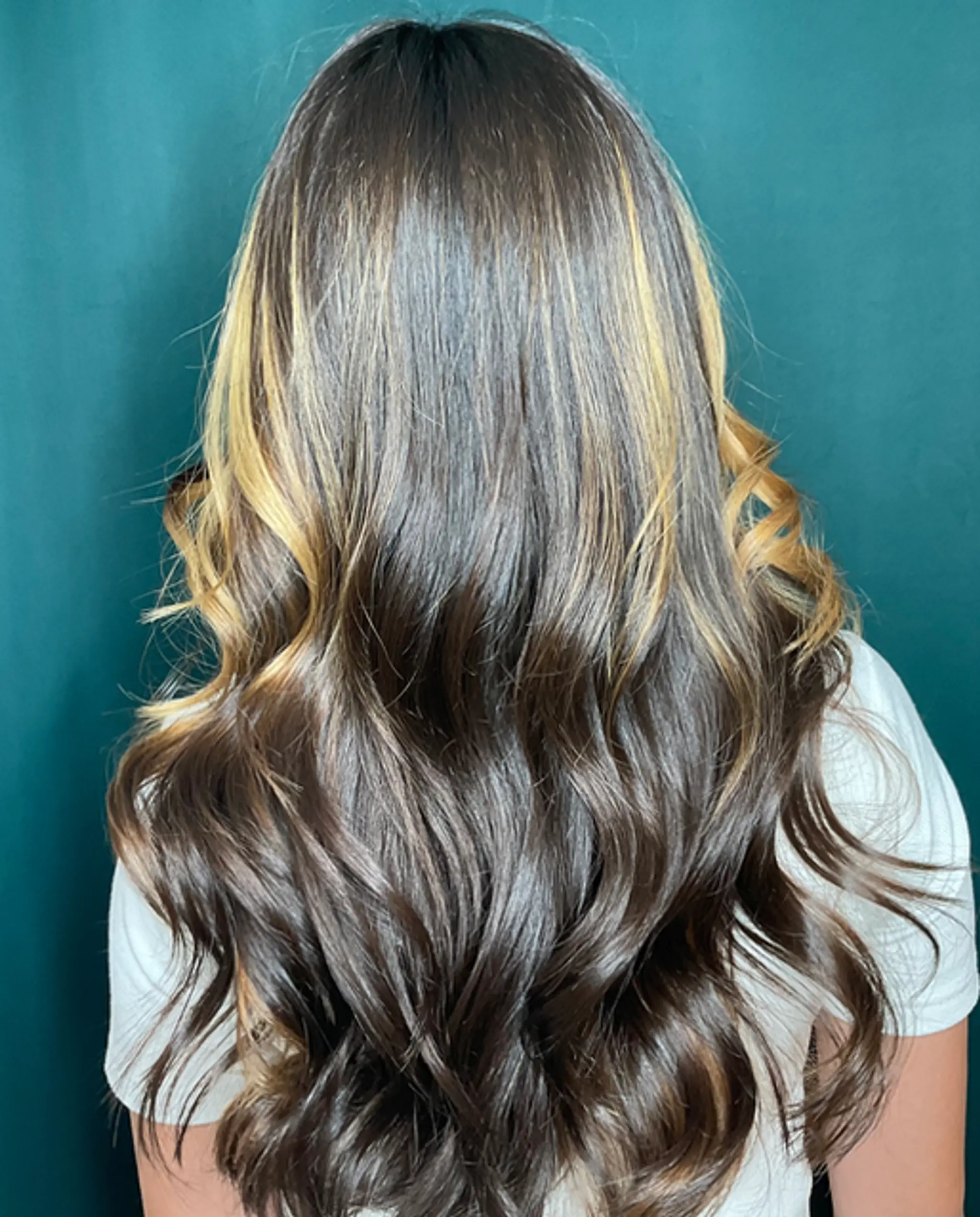 Long, cascading waves in a dark brown base with golden blonde highlights, featuring a soft and natural shine.