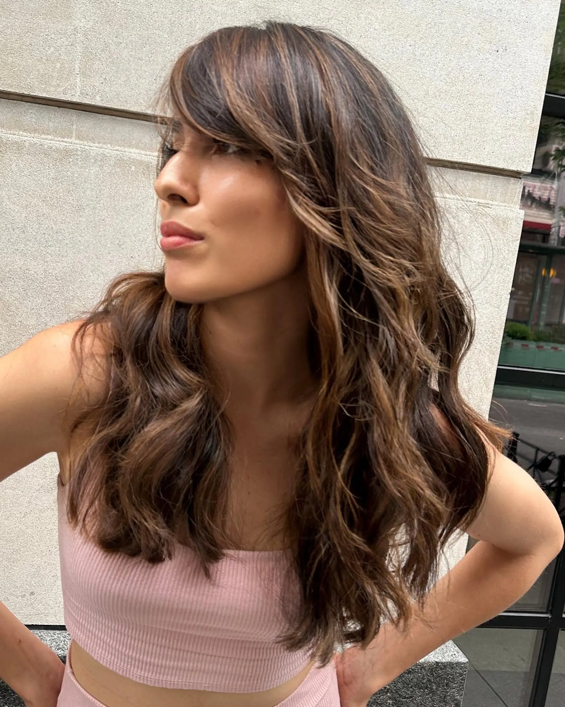 Long dark brown hair enhanced with warm caramel highlights, styled into tousled waves with layered side bangs. 1