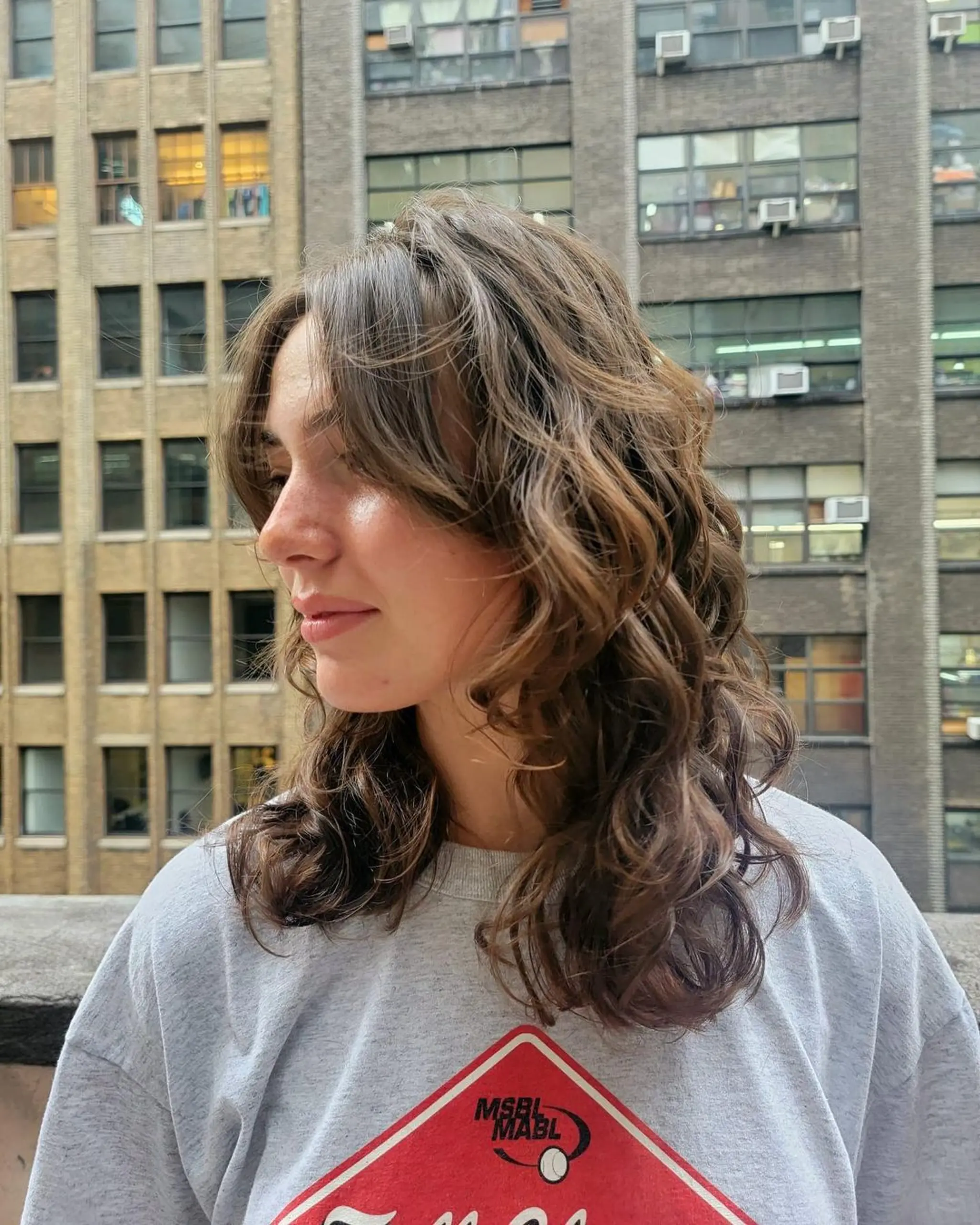 Medium-length, brown wavy hair with natural waves and loose curls, featuring layered cuts and a side parting. Subtle highlights add a sun-kissed look to the urban chic style.