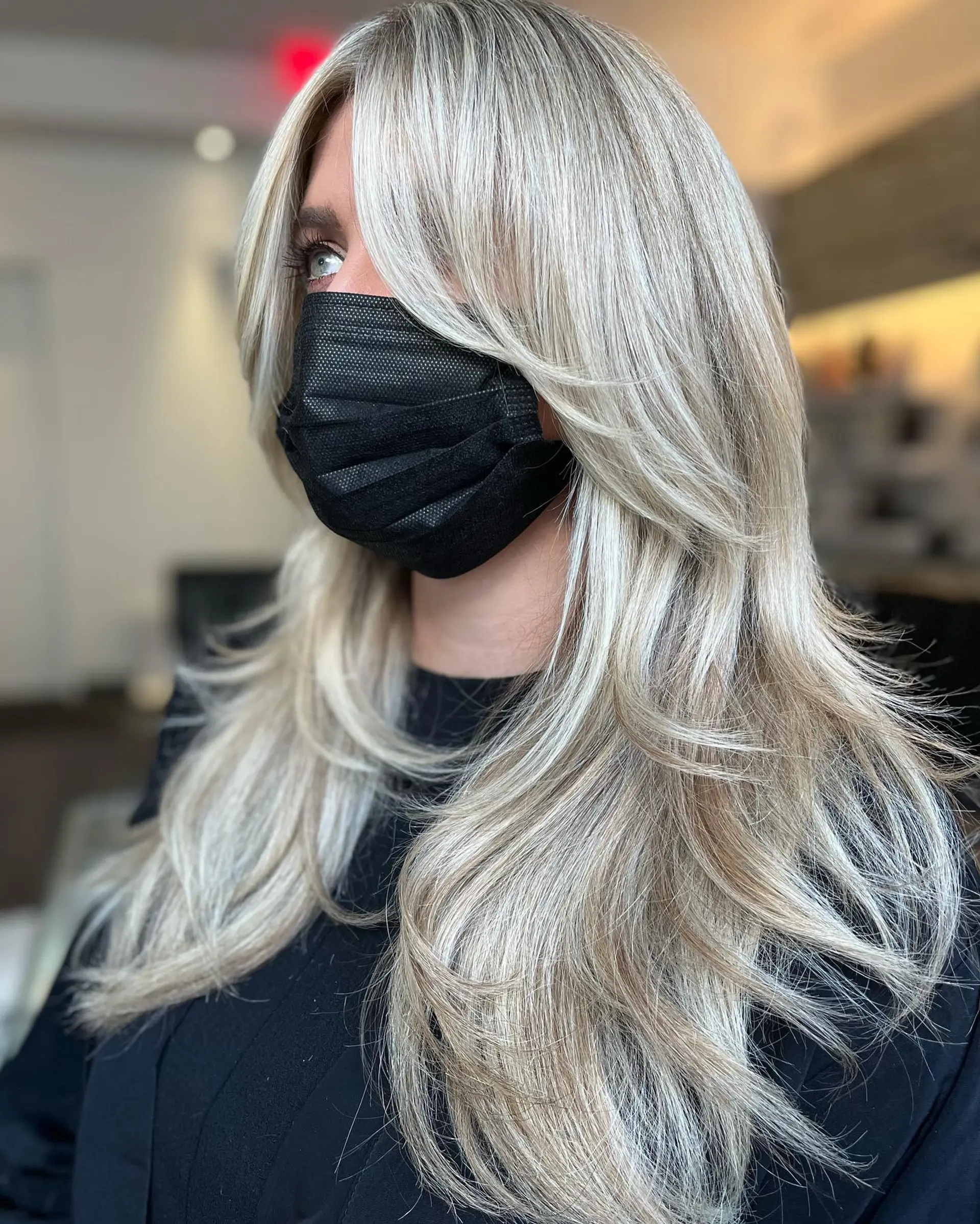 Long bright blonde hair styled with feathered face-framing layers and long curtain bangs, finished with a smooth blowout. 1