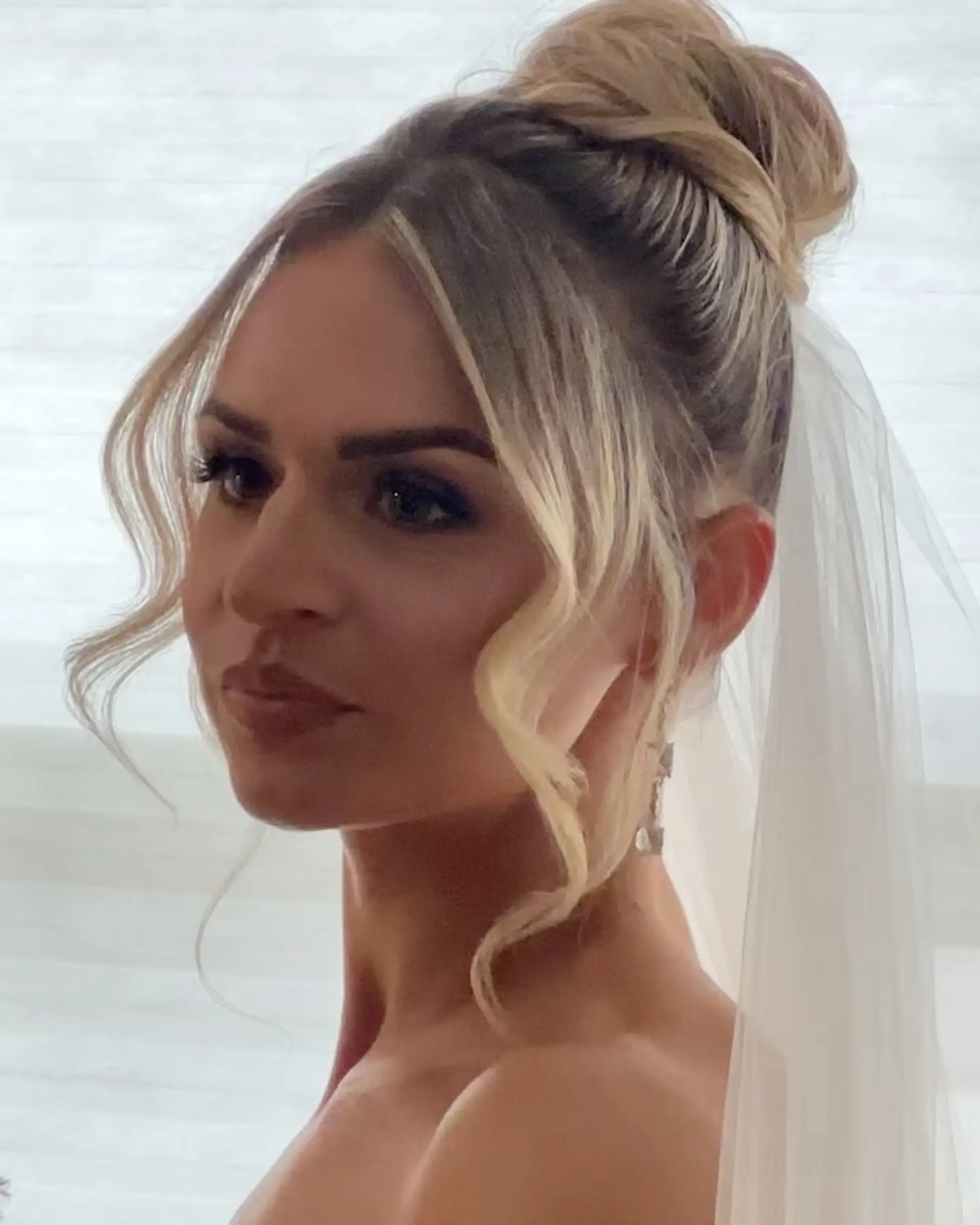 A sophisticated bridal high bun with softly curled face-framing tendrils and a delicate veil attachment. 1