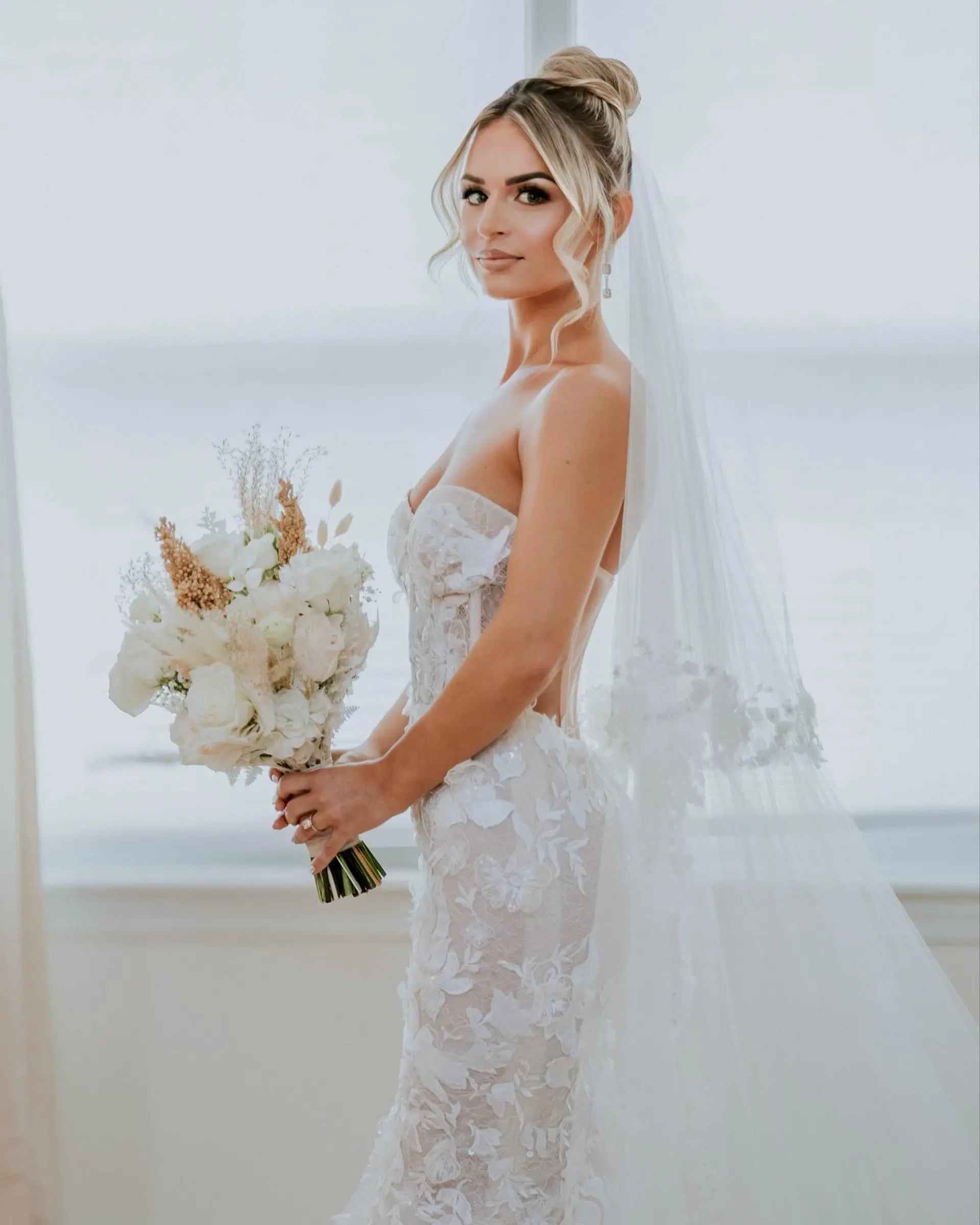 An elegant bridal high bun updo with romantic, soft face-framing wavy tendrils and dimensional blonde highlights. 1