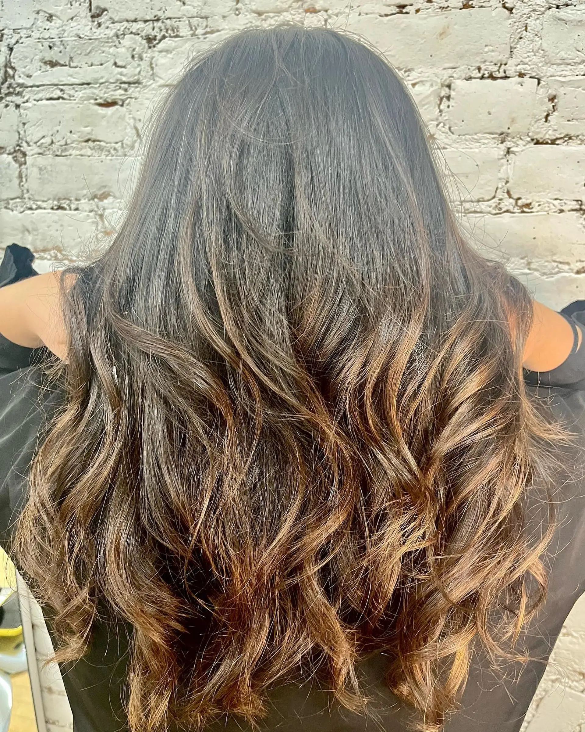 Long layered dark hair enhanced with warm caramel balayage and styled into loose waves. 1