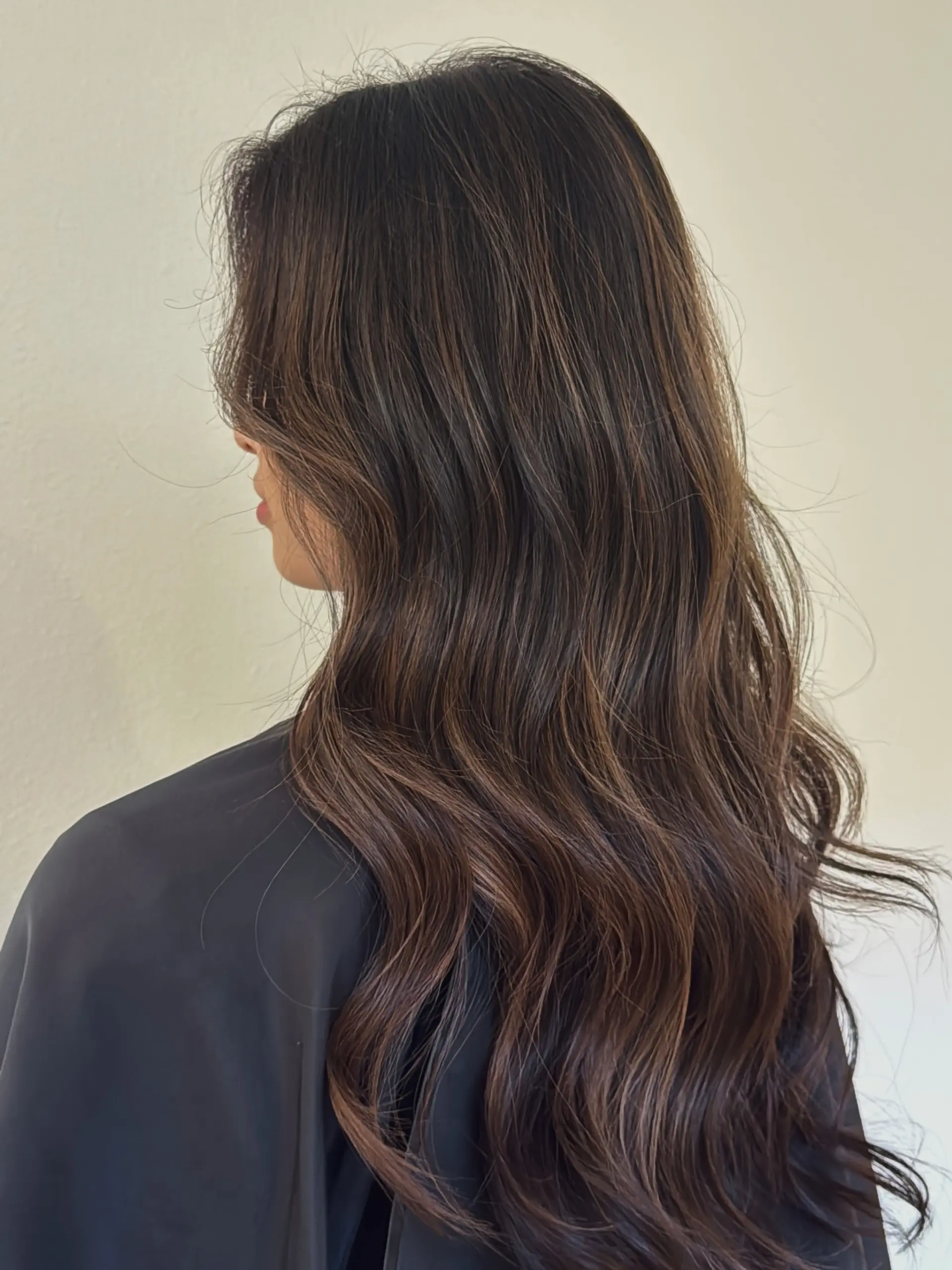 Long dark brown hair enhanced with subtle warm balayage highlights, styled in loose, natural waves. 1