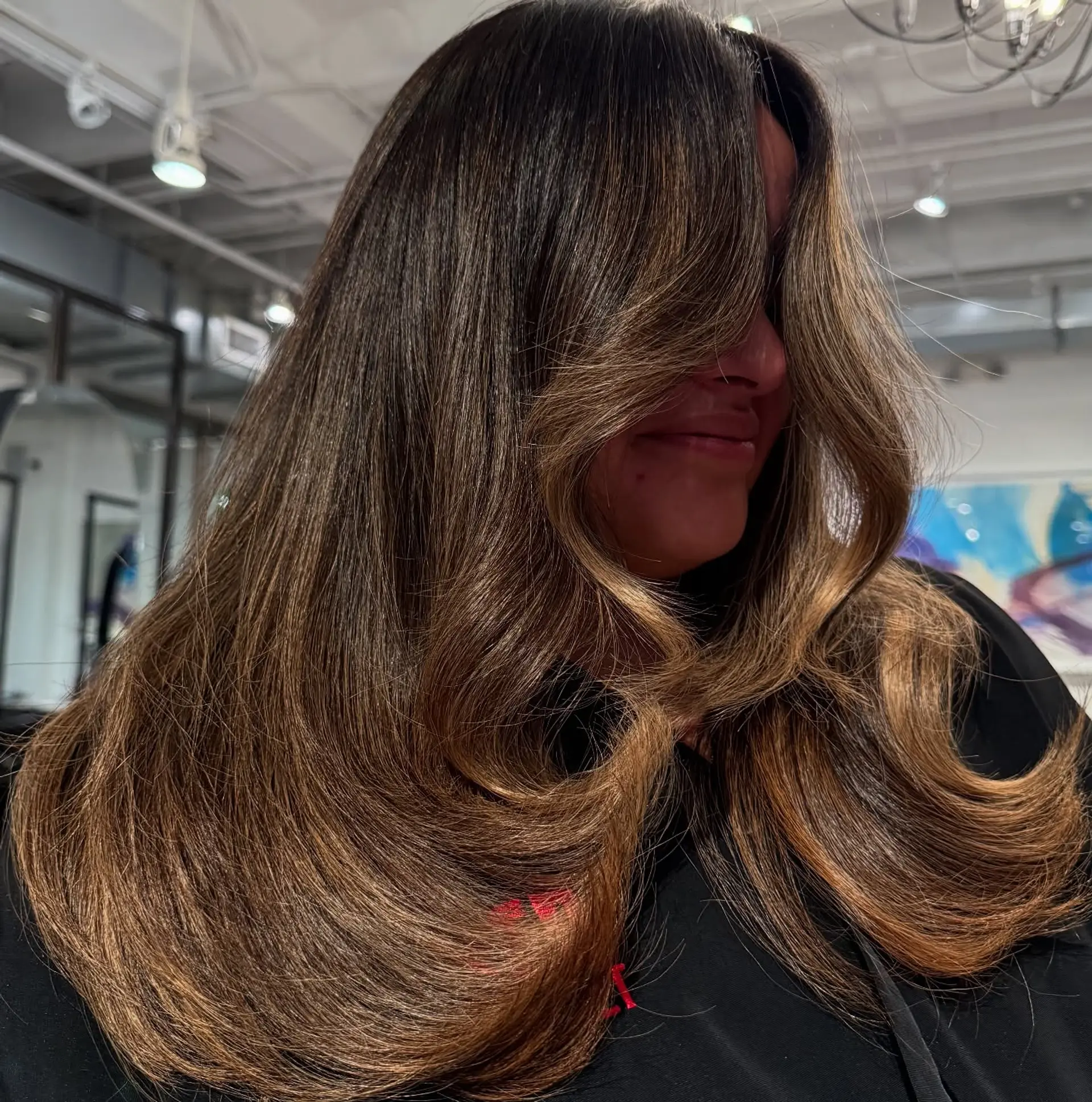 Long brunette hair enhanced with seamless golden caramel balayage highlights, styled into a smooth, voluminous blowout with rounded curved ends. 1