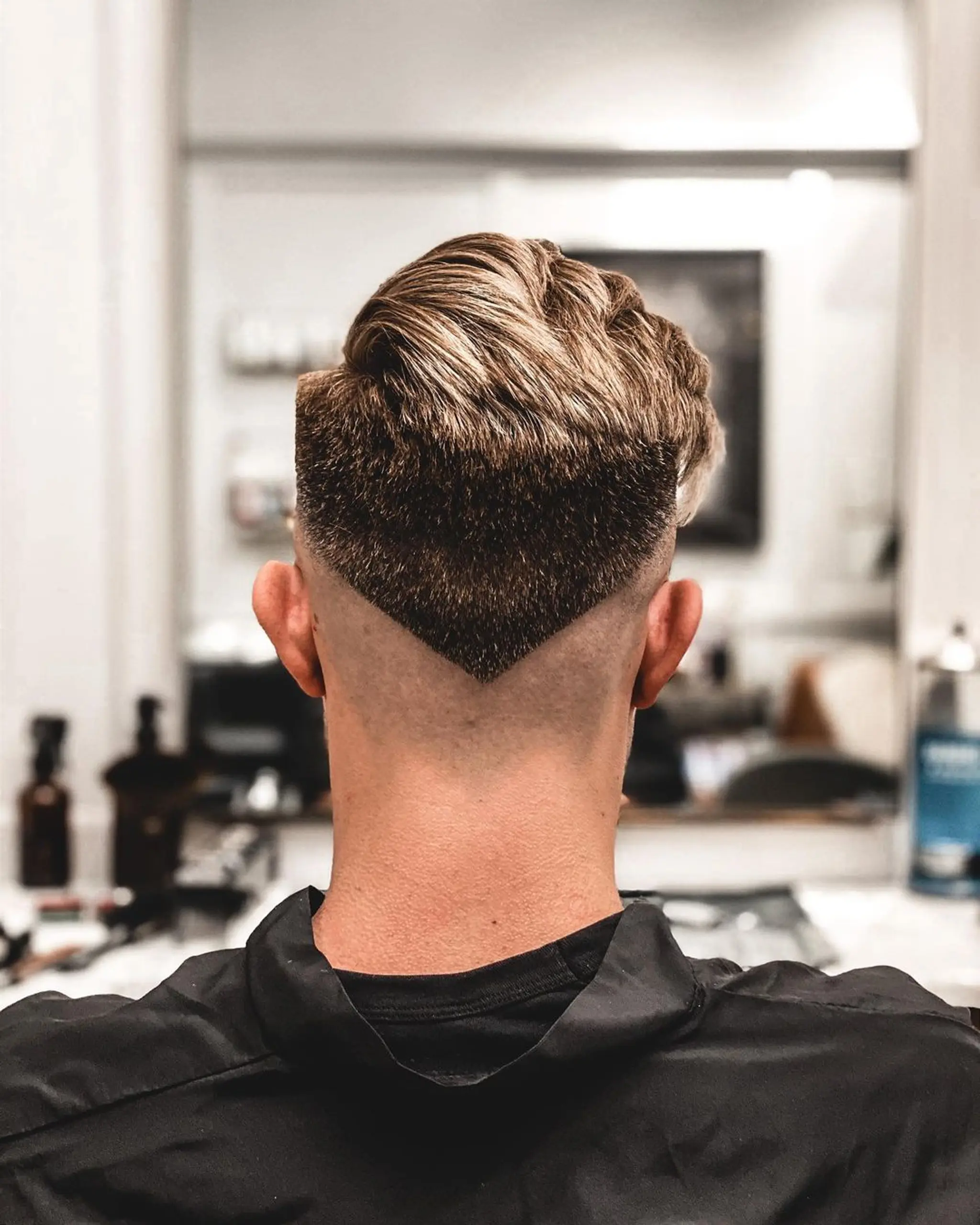The hairstyle features a high contrast fade with the lower section shaved into a sharp V shape. The top section is longer and voluminous, with blonde and brown highlights, styled in an edgy, modern manner.