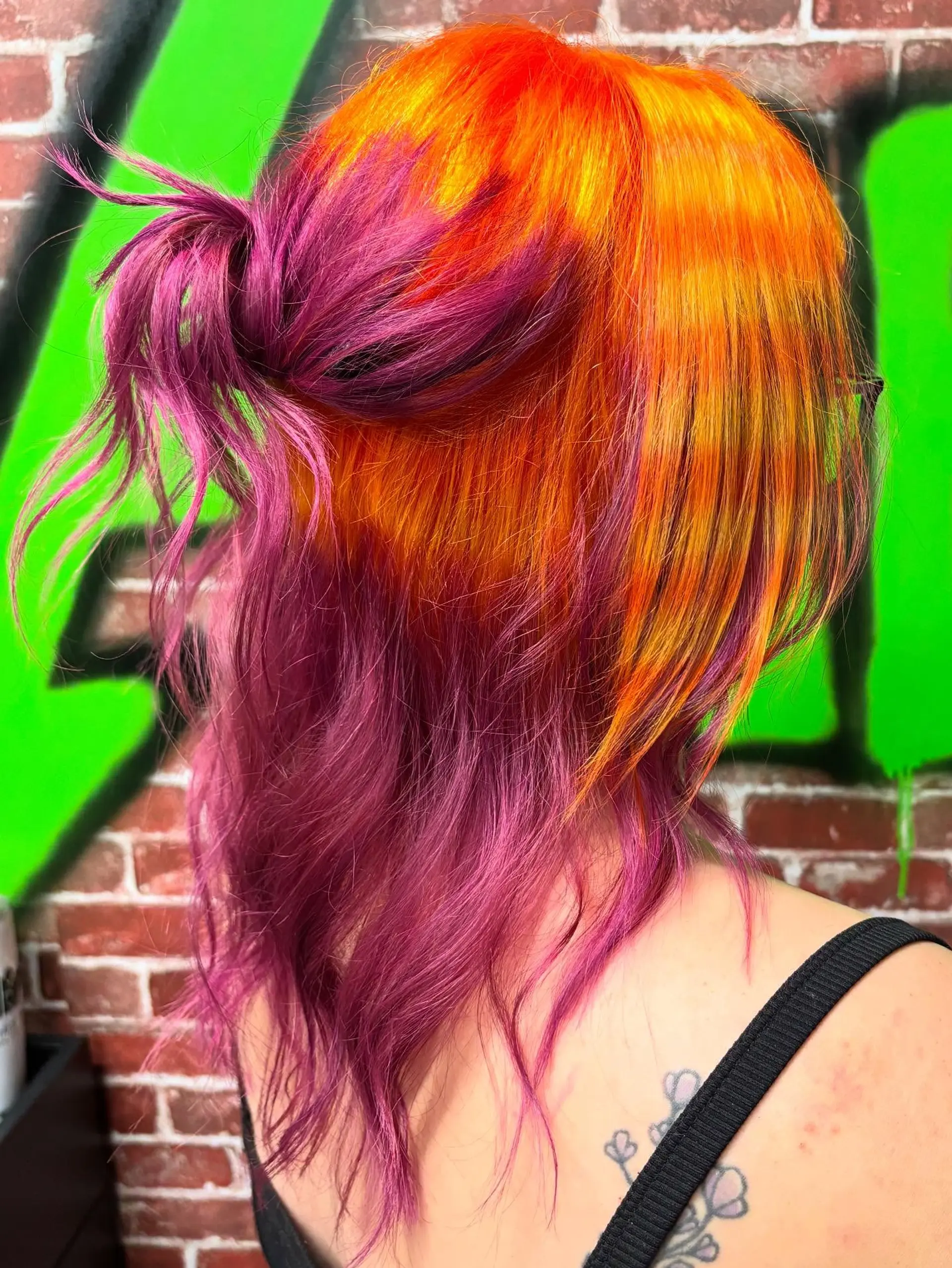 A vibrant split vivid color look featuring neon orange and yellow on top contrasted with rich magenta tones underneath, styled into a casual half-up bun. 1