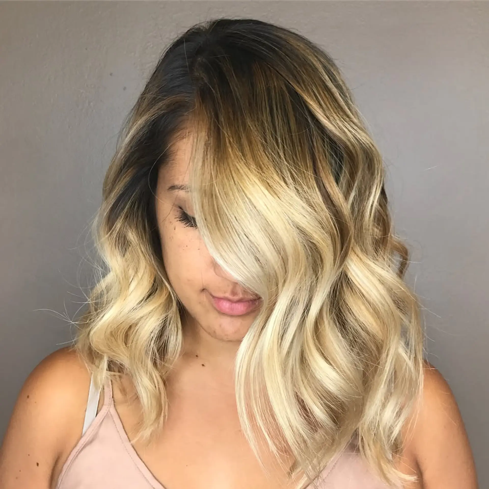A medium-length lob featuring a dark root melting into bright blonde balayage styled with soft, loose waves. 1