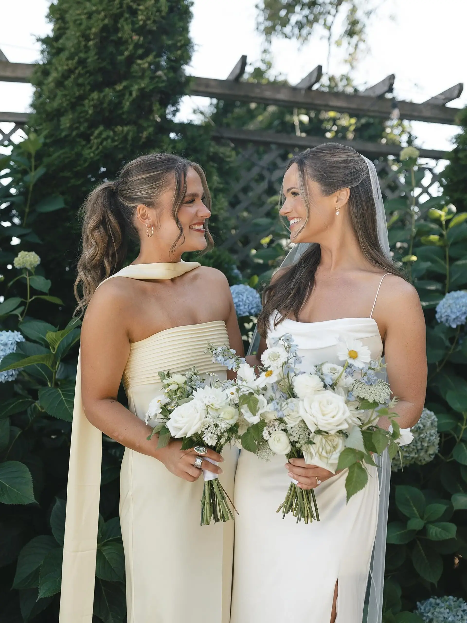Elegant bridal and bridesmaid hair styling featuring a wavy textured high ponytail with soft tendrils and a polished bridal half-up style paired with a veil. 1