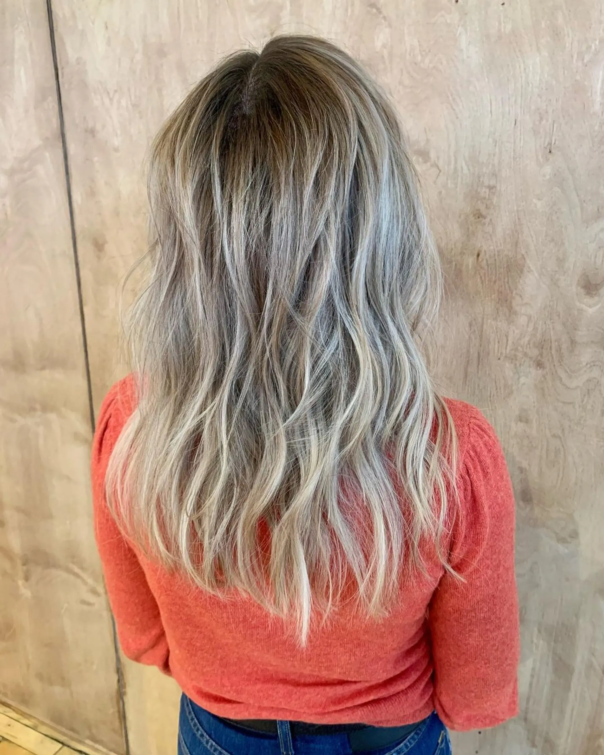 Long, wavy hairstyle with a balayage color transitioning from light brown roots to ash blonde ends, featuring natural, textured waves and a root shadow for added depth.