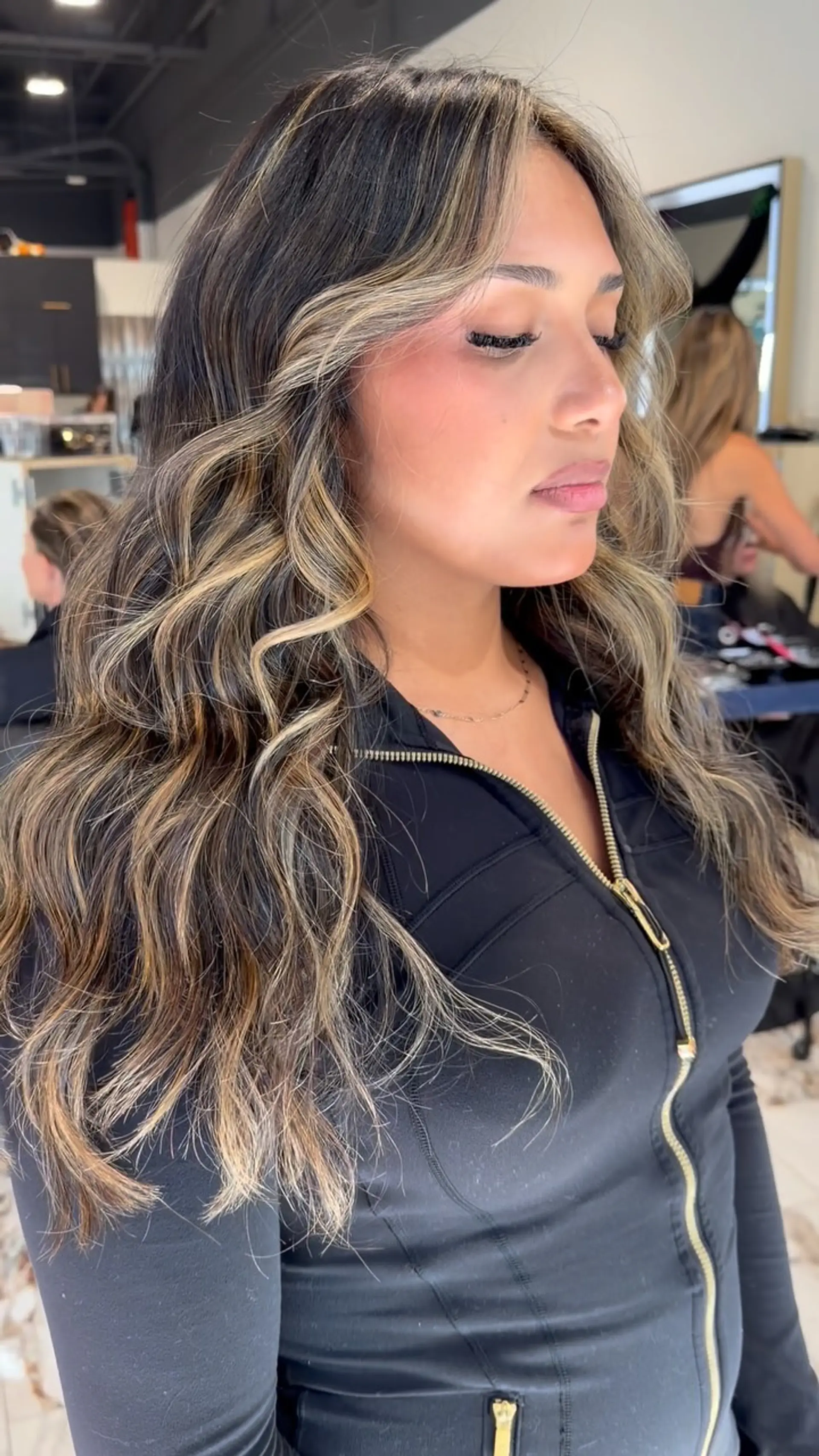 Long brunette hair styled in soft, loose waves featuring warm caramel and blonde balayage highlights with face-framing pieces. 1
